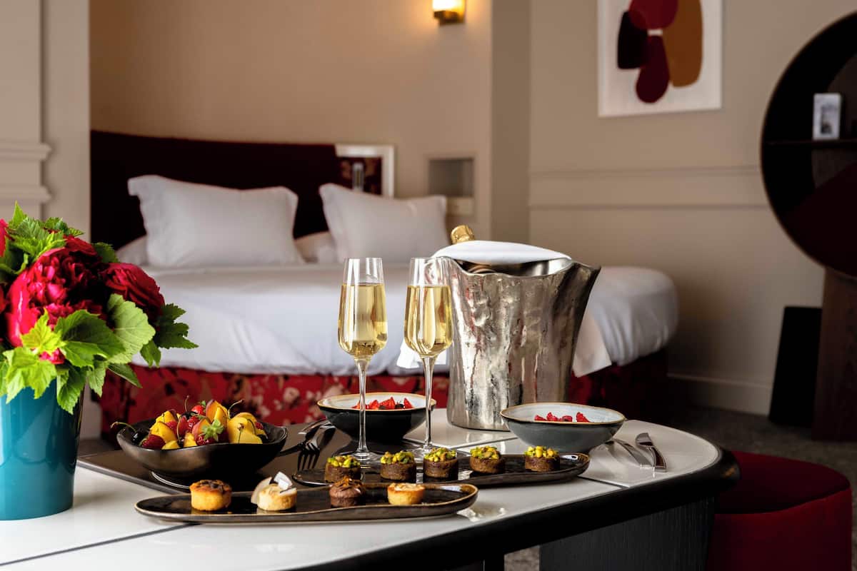 A dining table with a variety of food items, two glasses of champagne, a vase with flowers, and a bed in the background.