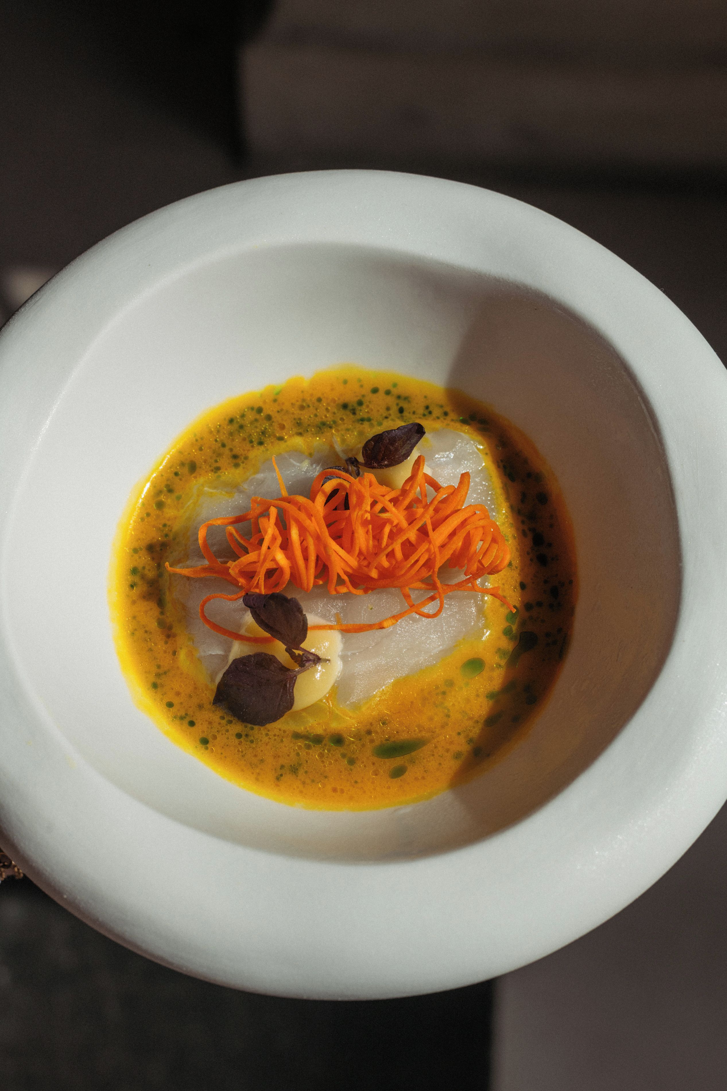 A white bowl containing a dish with a yellow sauce, shredded carrots, and a piece of fish.