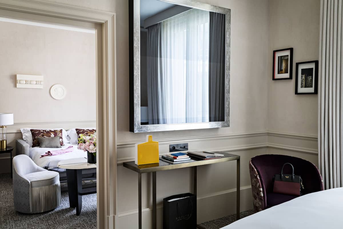 A hotel room with a bed, a desk with a lamp, a chair, a small table, and a mirror.