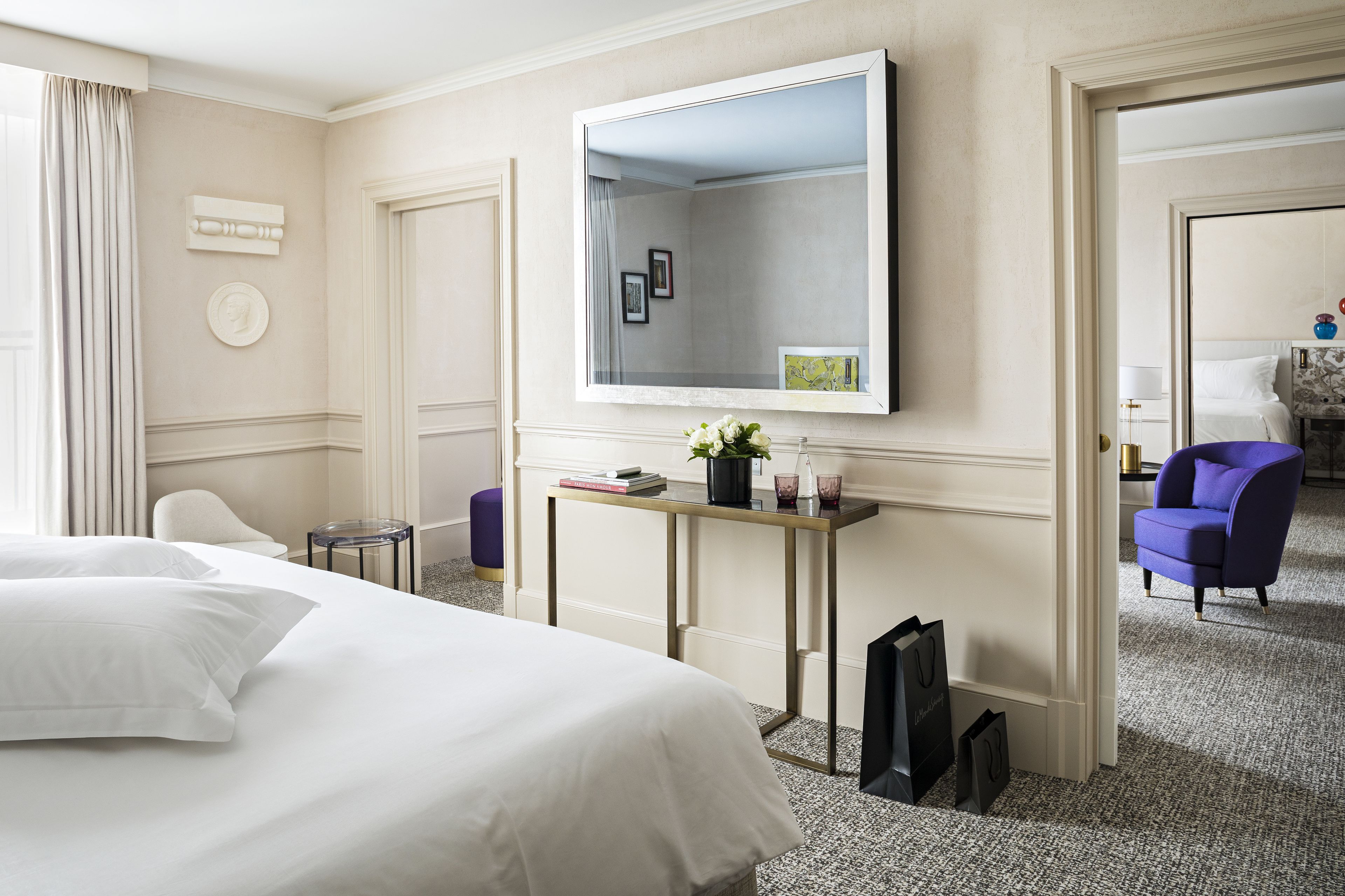 Habitación de hotel con una cama grande, una mesita con un florero y libros, un espejo y un sillón morado.
