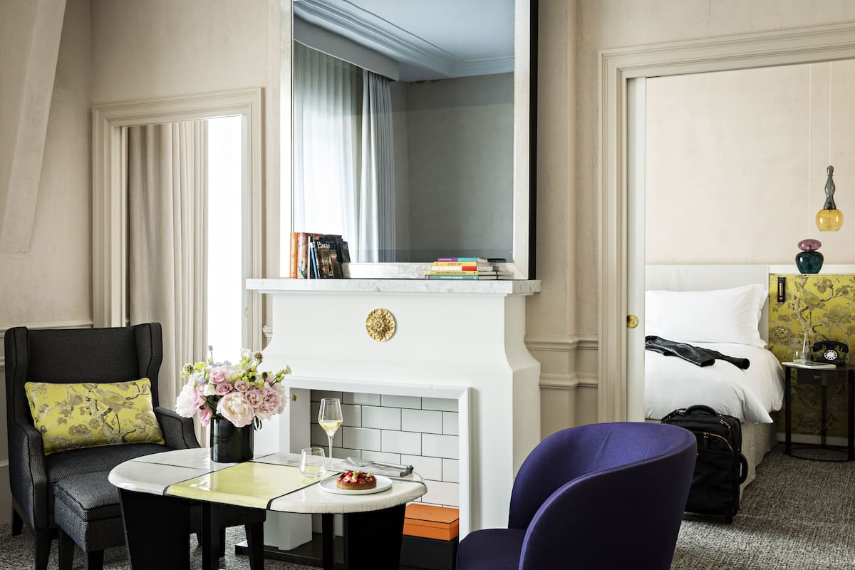 A hotel room with a dining table, a chair, a sofa, a bed, a nightstand, and a fireplace.