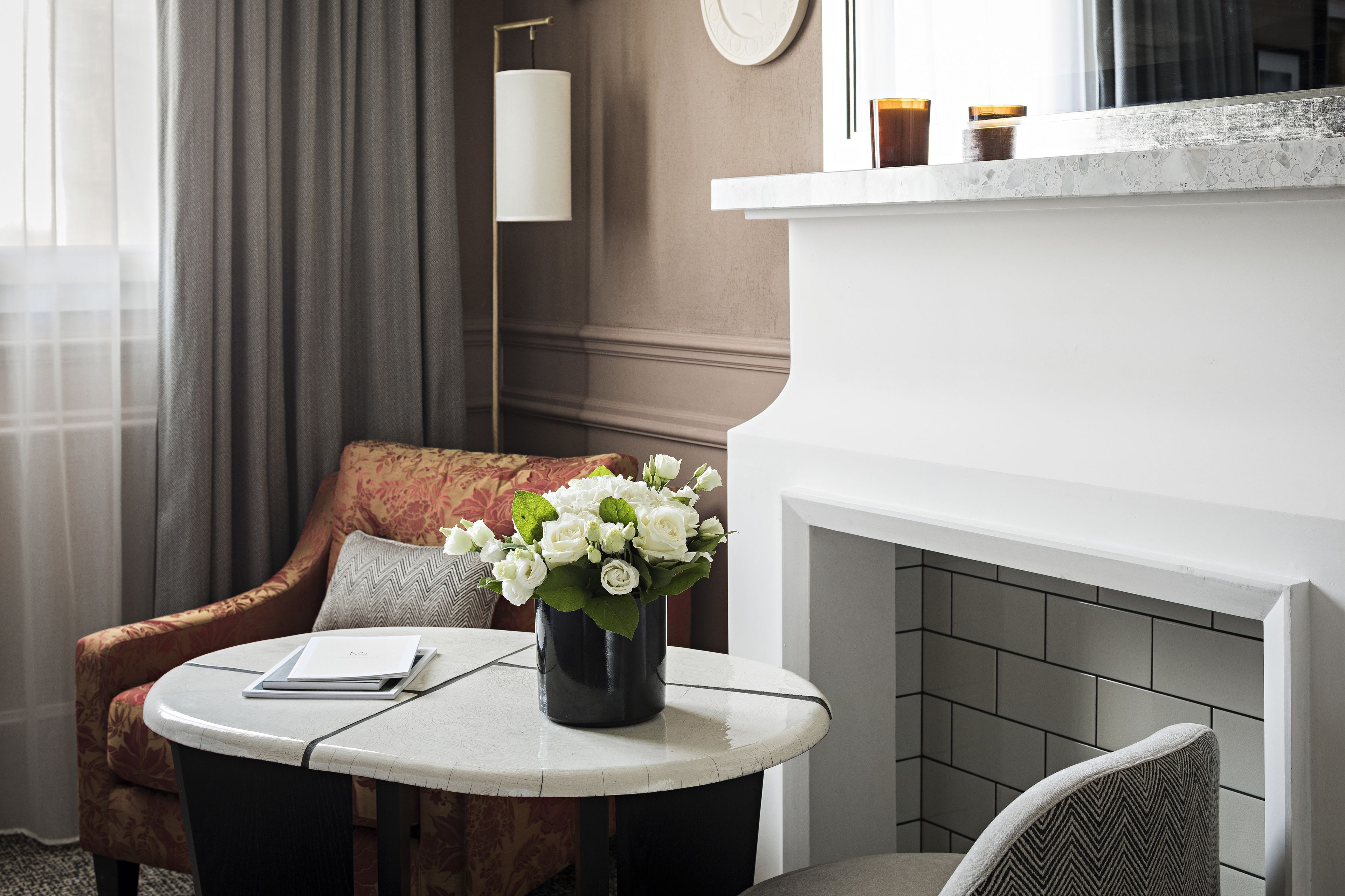 A hotel room with a round table, a sofa, a vase of white flowers, a fireplace, and a clock on the wall.