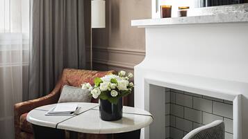 A hotel room with a round table, a sofa, a vase of white flowers, a fireplace, and a clock on the wall.