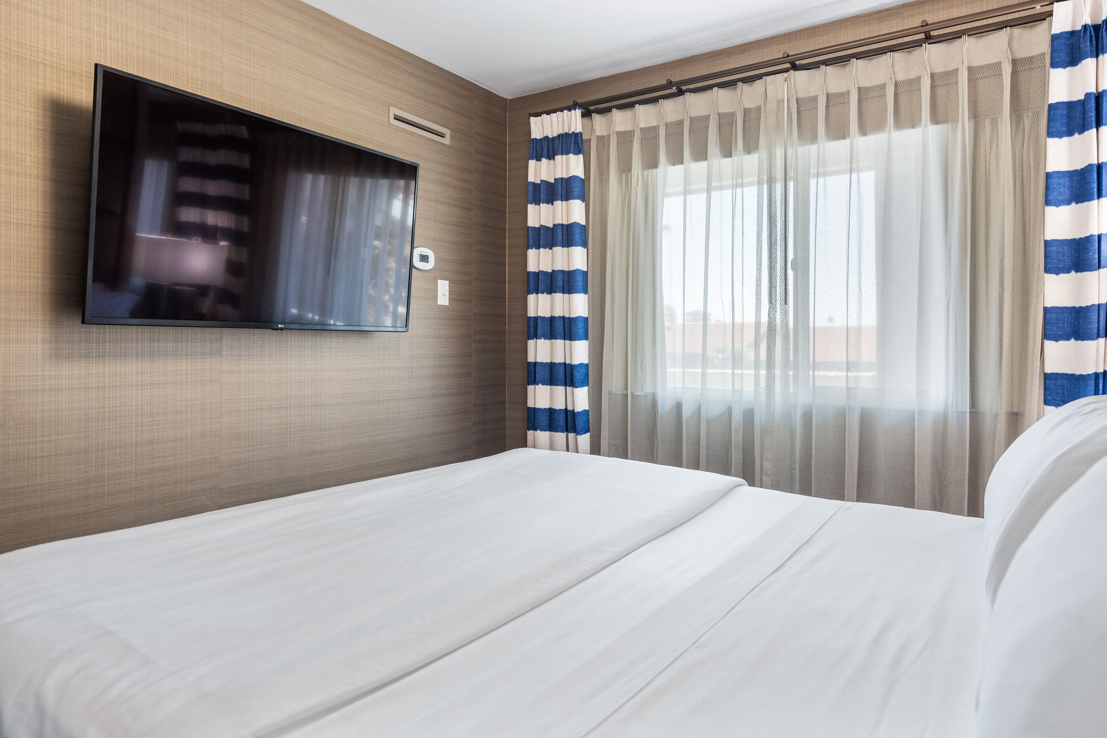 A hotel room with a flat-screen TV mounted on the wall, a bed with white bedding, and a window with striped curtains.