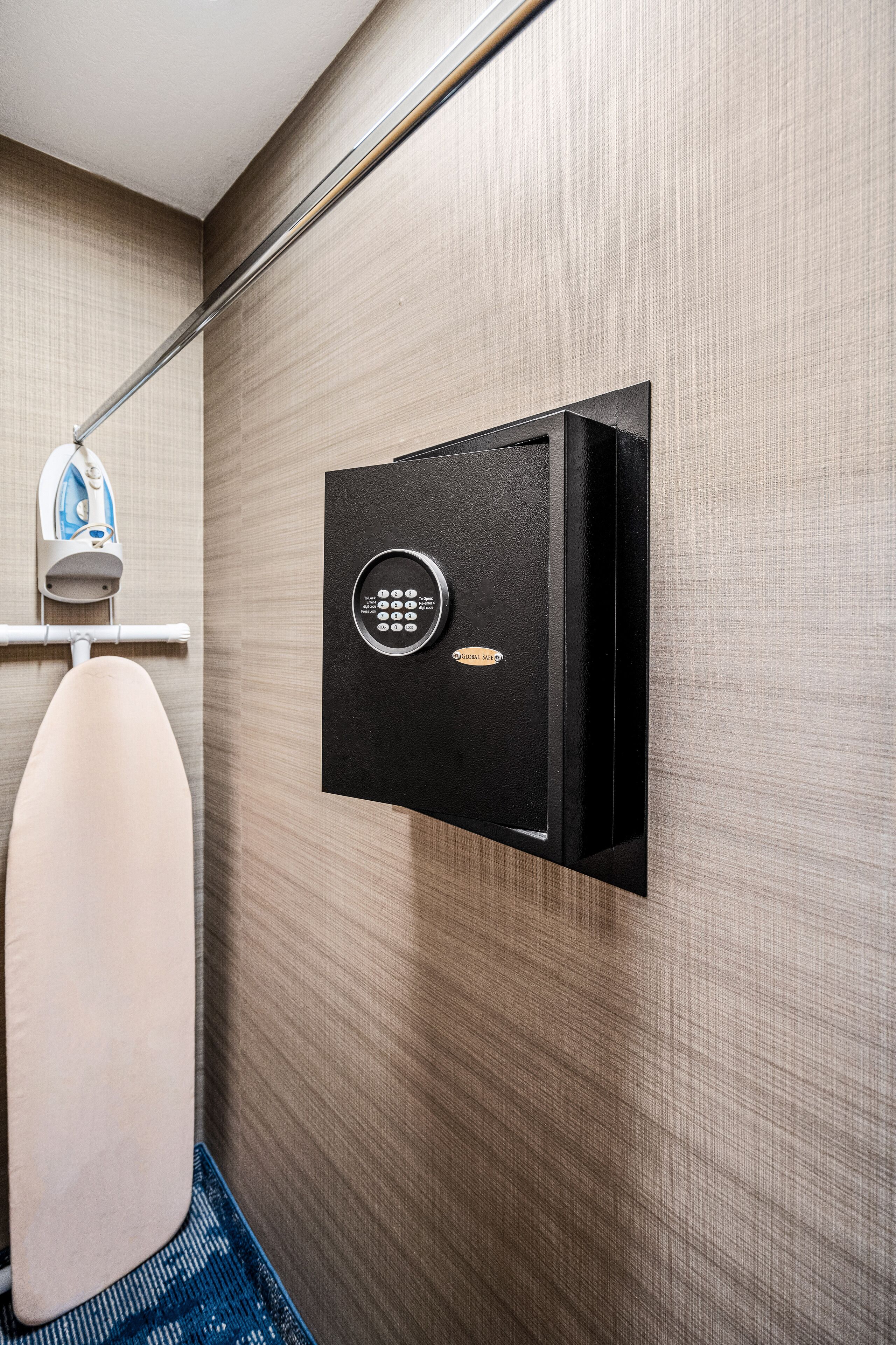A wall-mounted safe with a keypad lock, a hanging iron, and a towel rack in a hotel room.