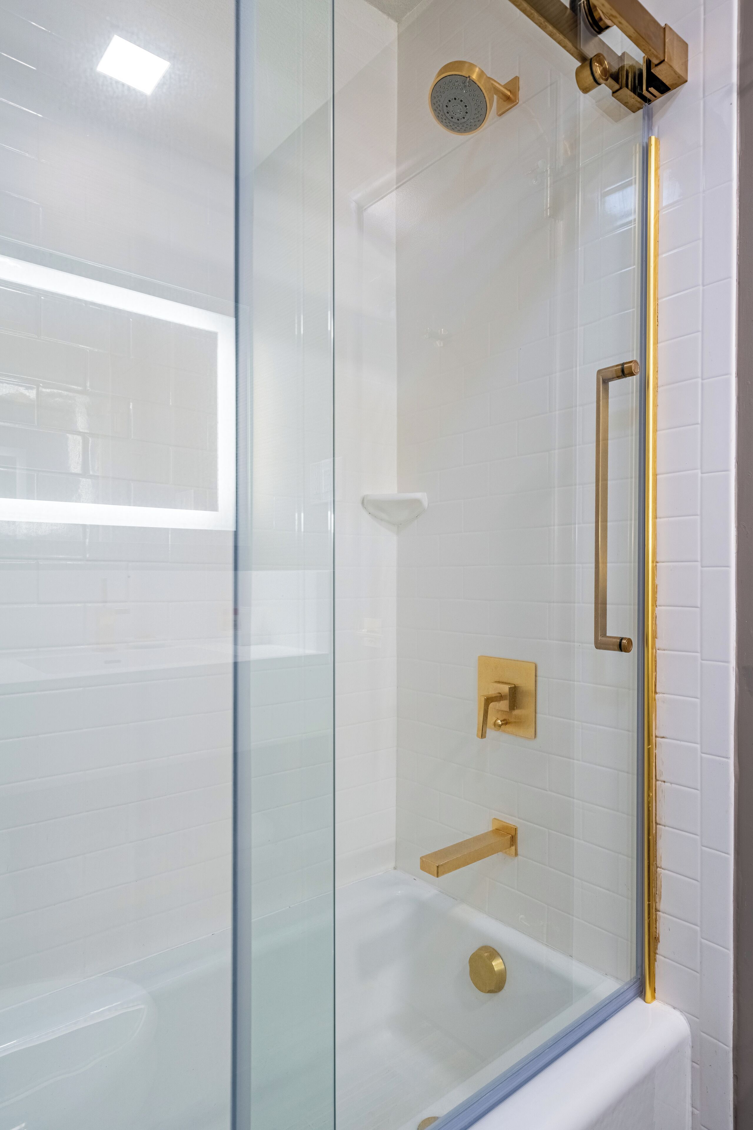 A glass-enclosed shower with a gold faucet and showerhead, white tiled walls, and a visible window.