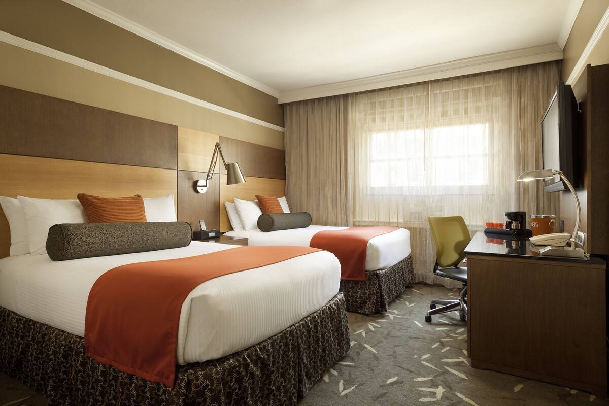 A hotel room with two beds, a desk, a chair, a TV, and a window with curtains.