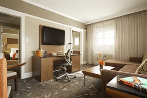 A hotel room with a desk, chair, sofa, and a TV mounted on the wall.