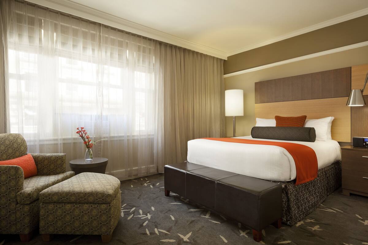 A hotel room with a bed, a chair, a small table with a vase, and a lamp.
