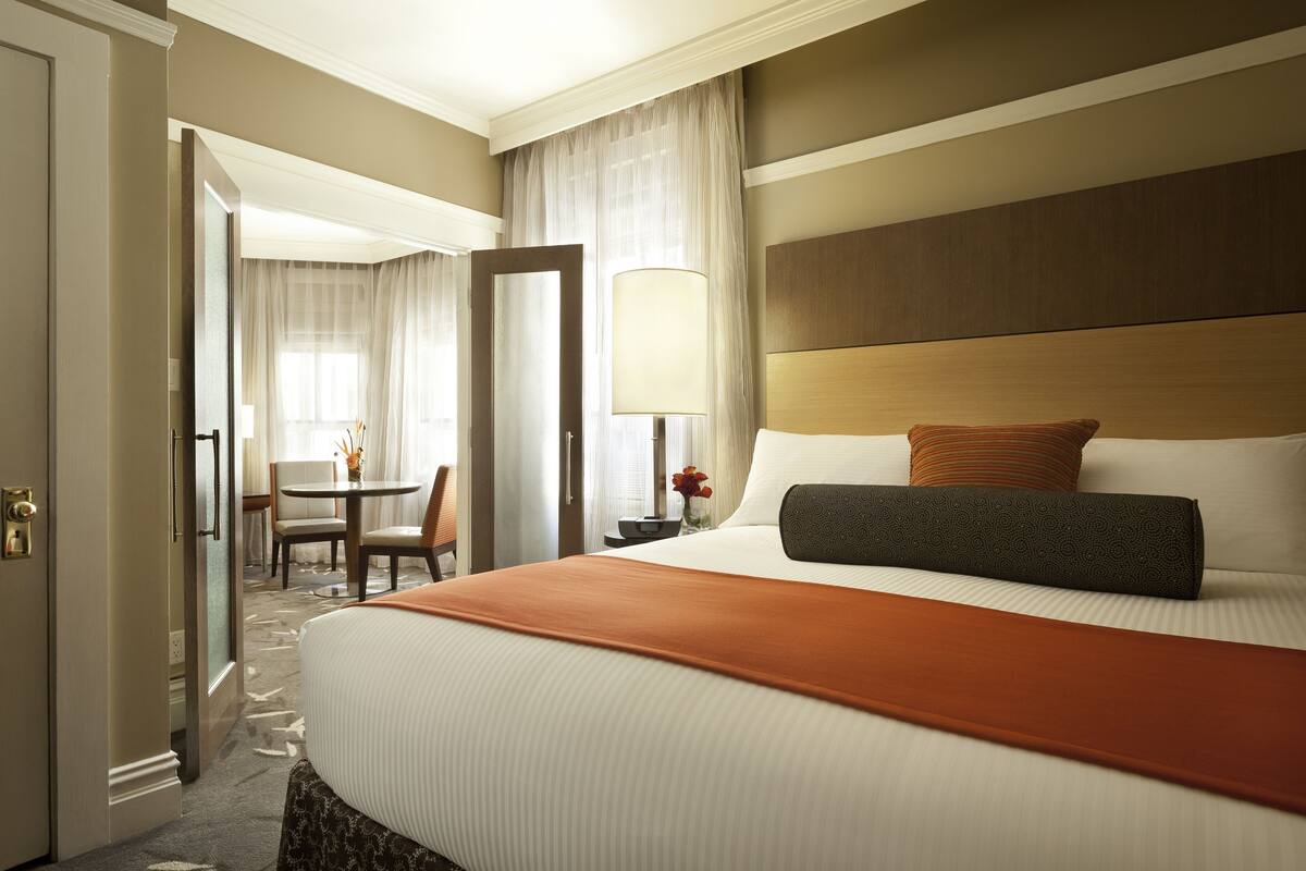A hotel room with a large bed, a bedside table with a lamp, a chair, and a desk with a vase of flowers.
