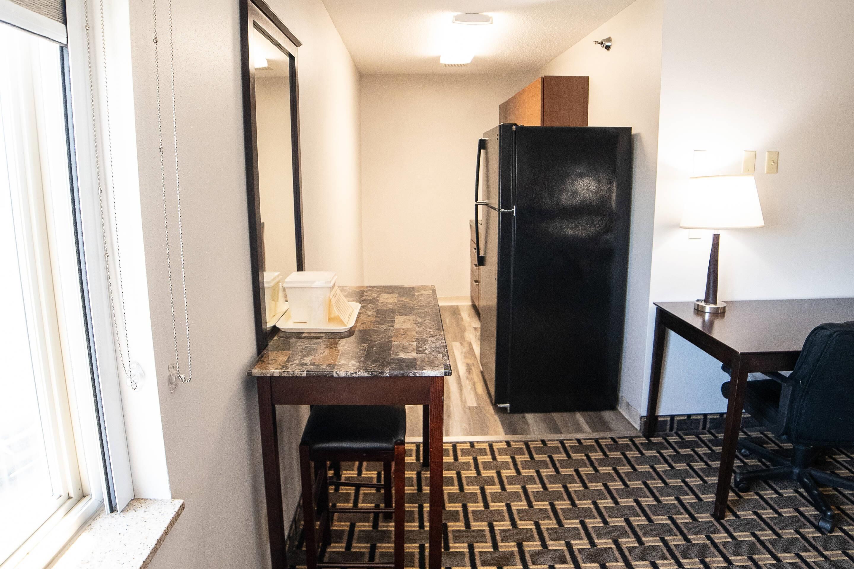 Une chambre d’hôtel compacte comprenant un coin cuisine, une petite table à manger, une chaise, un réfrigérateur et une lampe.
