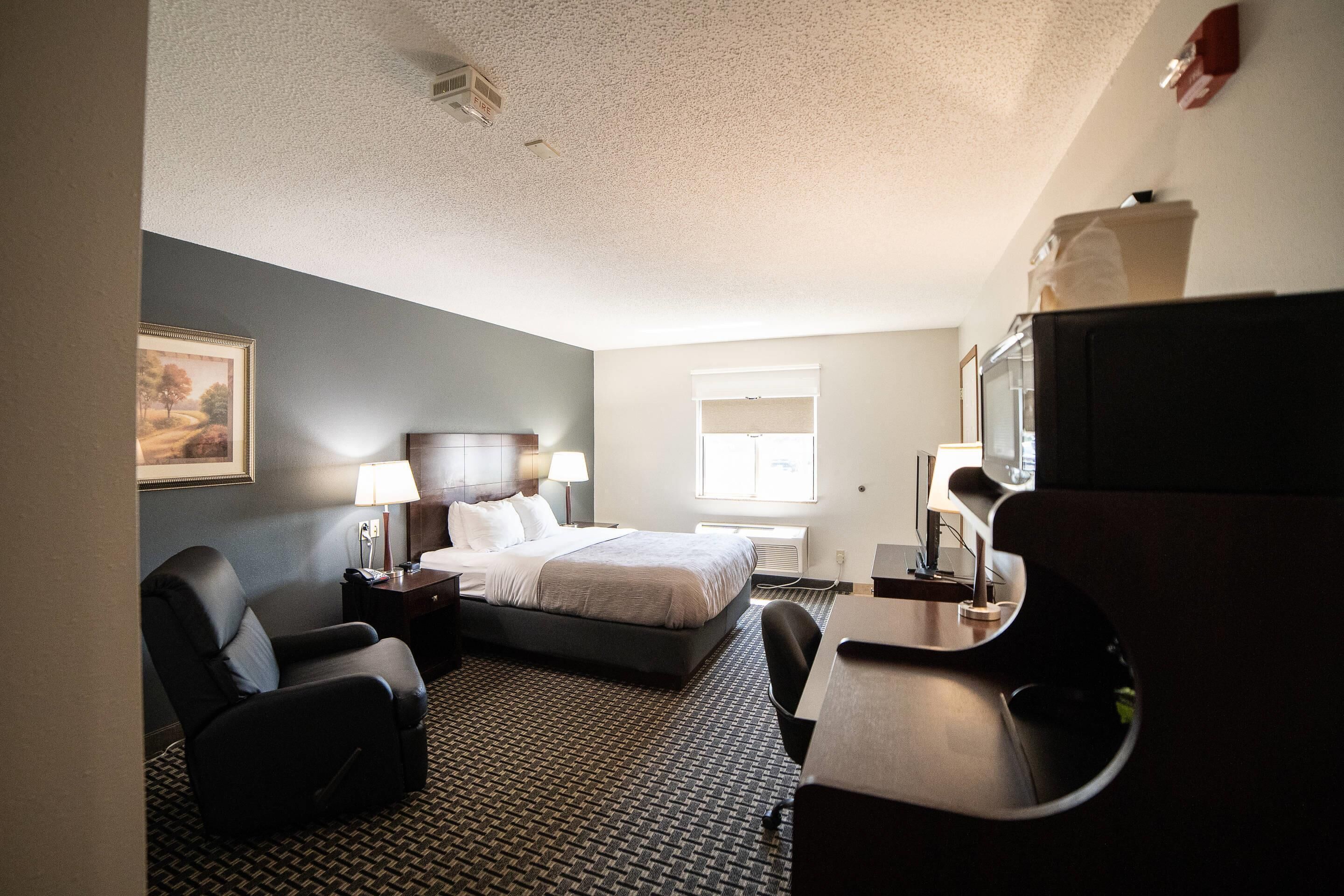A hotel room with a bed, two chairs, a desk, a television, a window, and a painting on the wall.