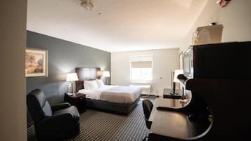 A hotel room with a bed, two chairs, a desk, a television, a window, and a painting on the wall.