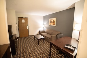 Una habitación de hotel con alfombra estampada, un sofá, una mesita, un escritorio con teléfono, una lámpara y un cuadro en la pared.