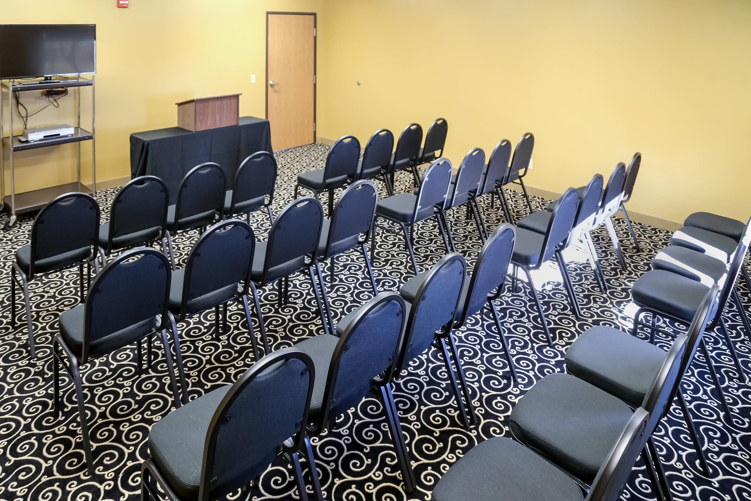 Une salle de conférence avec des rangées de chaises, un pupitre et un téléviseur posé sur un support.