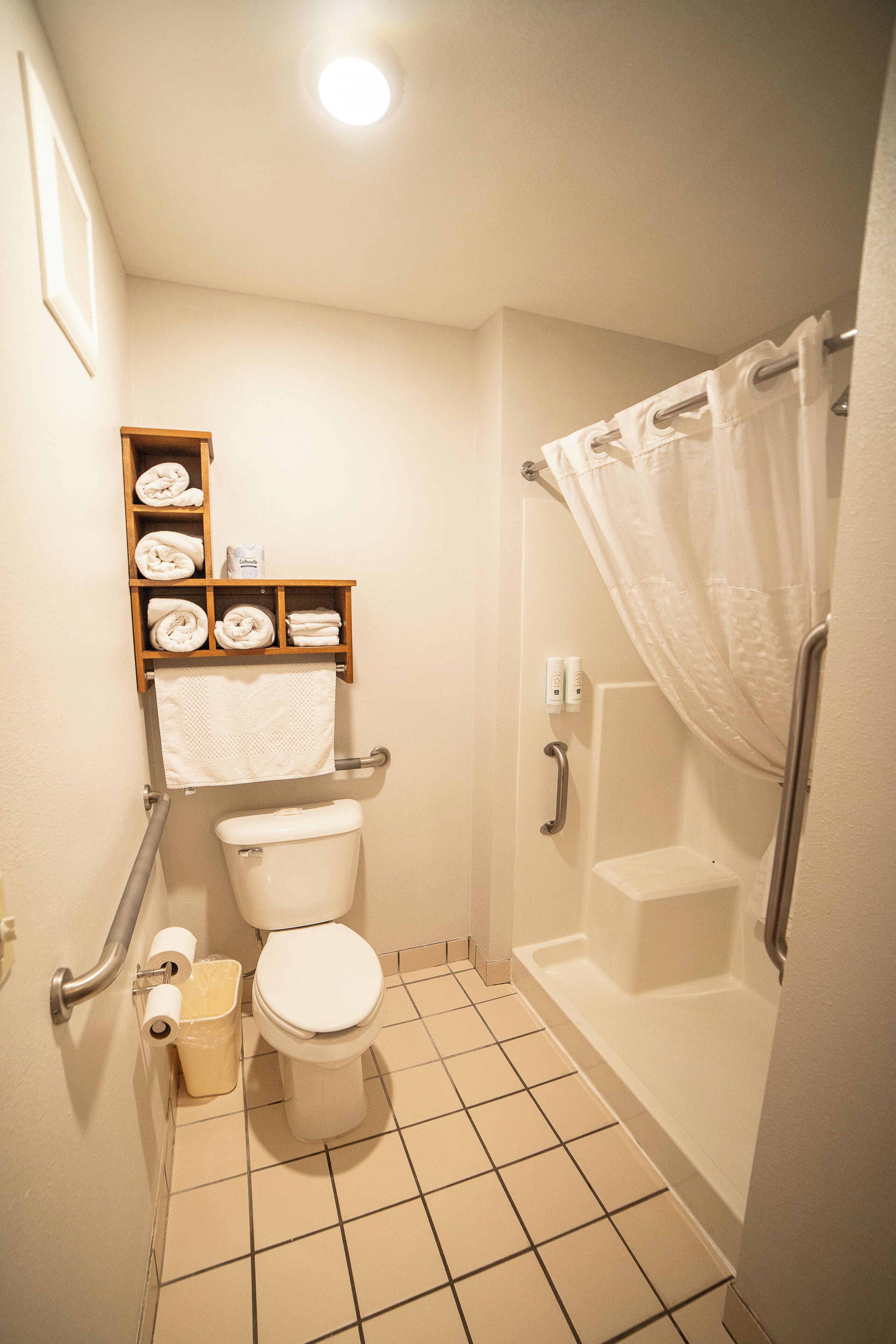 A small bathroom with a toilet, a shower with a curtain, a shelf with towels, and a handrail.