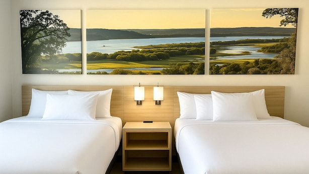 Habitación de hotel con dos camas, una mesita de noche con lámparas y un cuadro grande con un paisaje sobre las camas.
