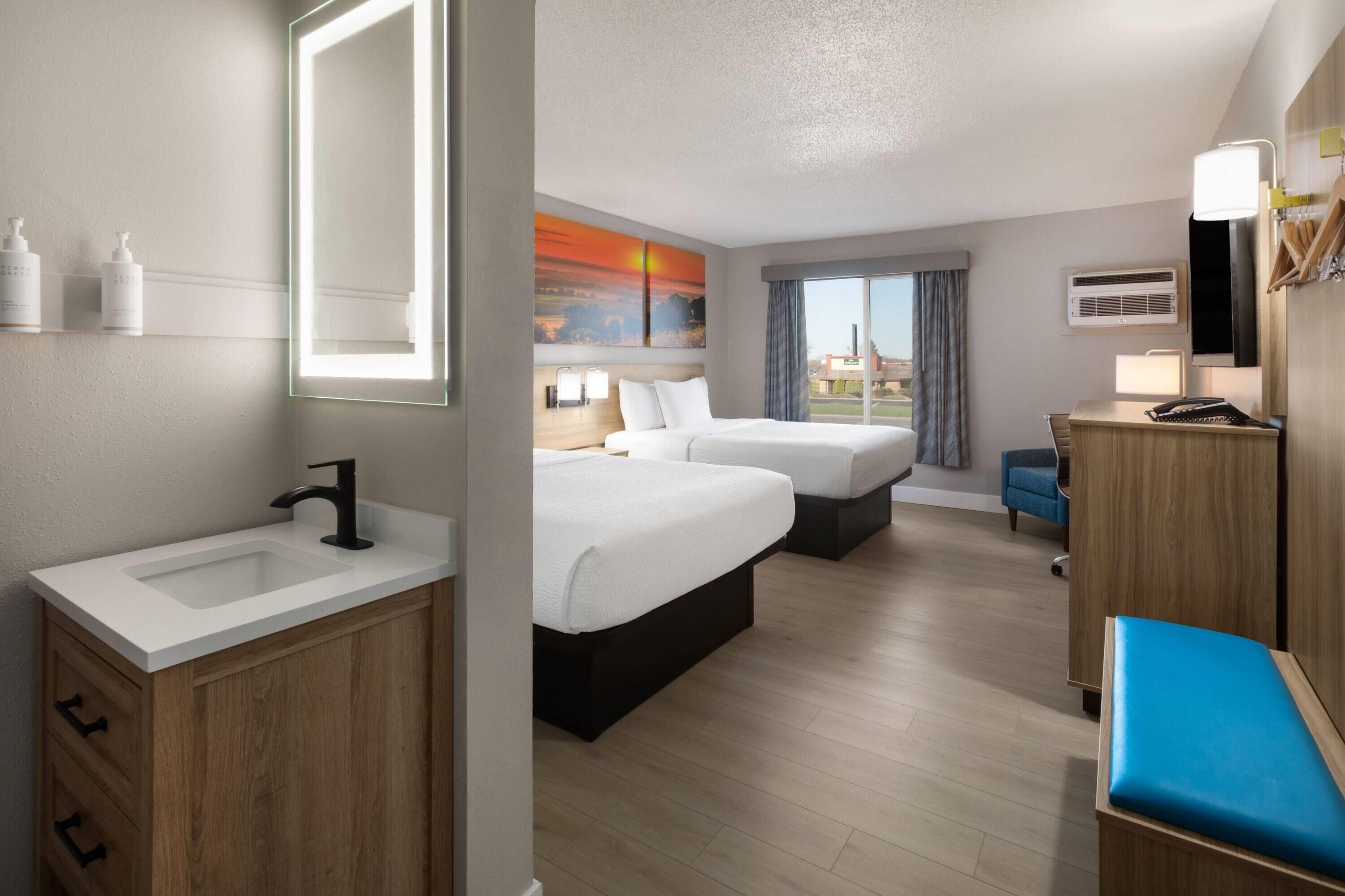 A hotel room with a large bed, a smaller bed, a desk, a chair, a bathroom with a sink and mirror, and a painting on the wall.