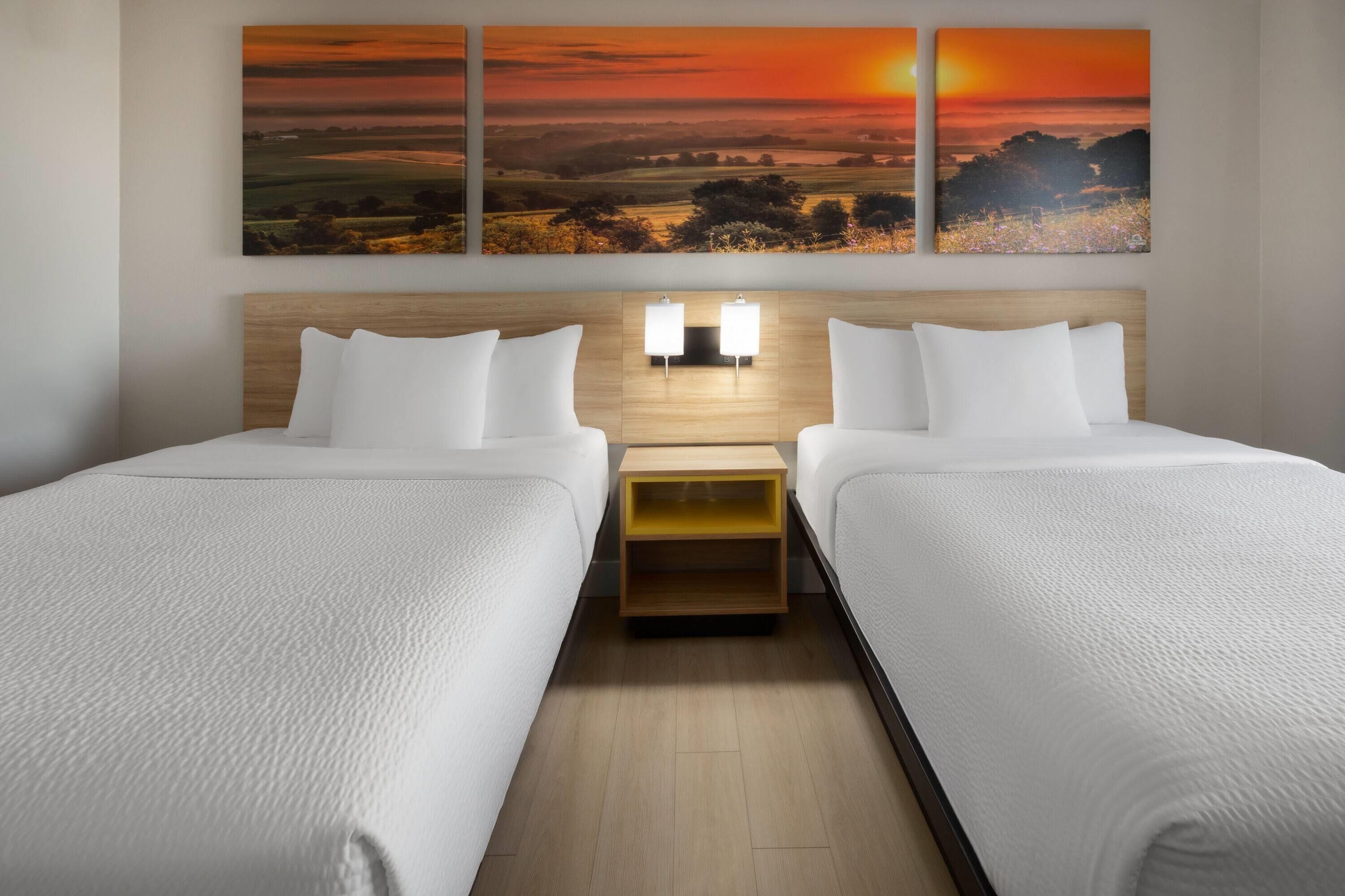 Habitación de hotel con dos camas, cabecero de madera, lámparas de noche y un cuadro con atardecer.