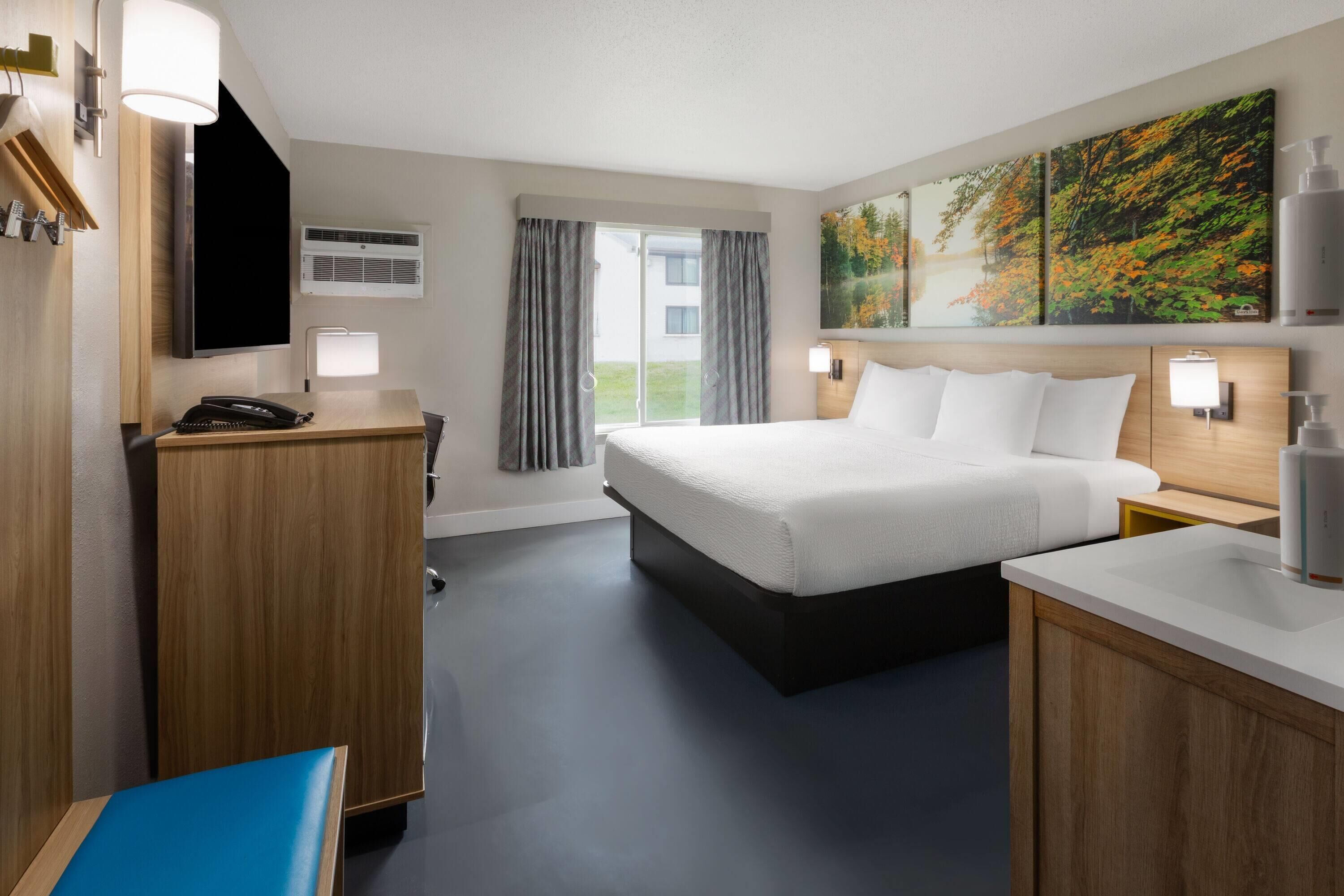 A hotel room with a bed, bedside tables, a desk, a chair, a TV, a painting, and a window with curtains.