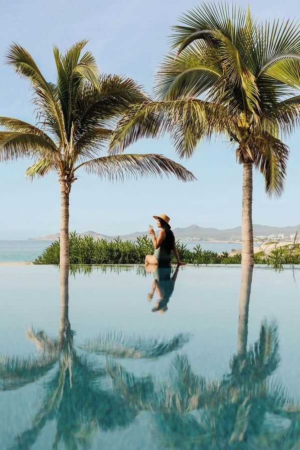 A person sitting on an infinity pool with palm trees and a view of the ocean.