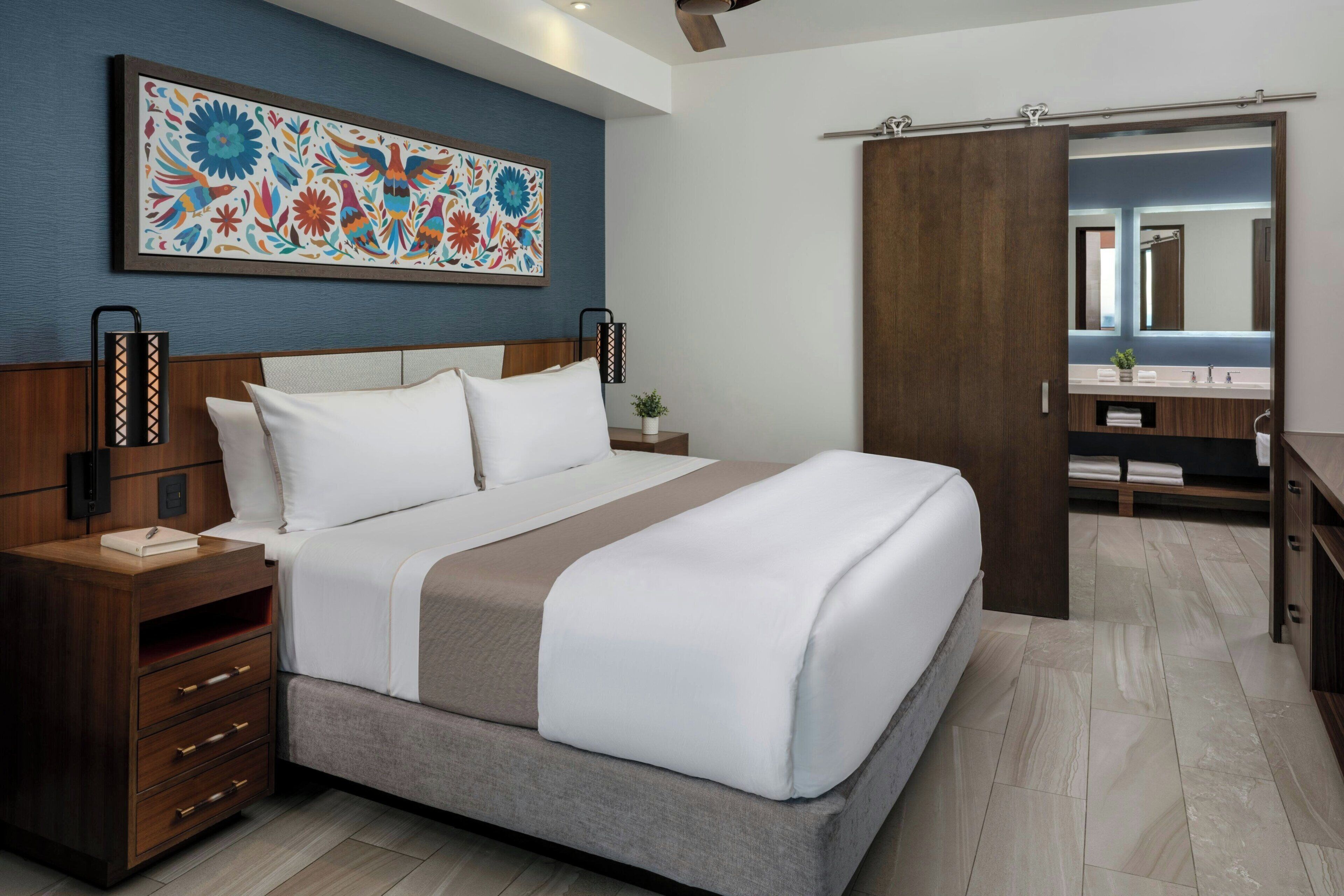 A modern hotel room with a large bed, a nightstand, a bedside table, a wall-mounted lamp, a mirror, and a door leading to a bathroom.