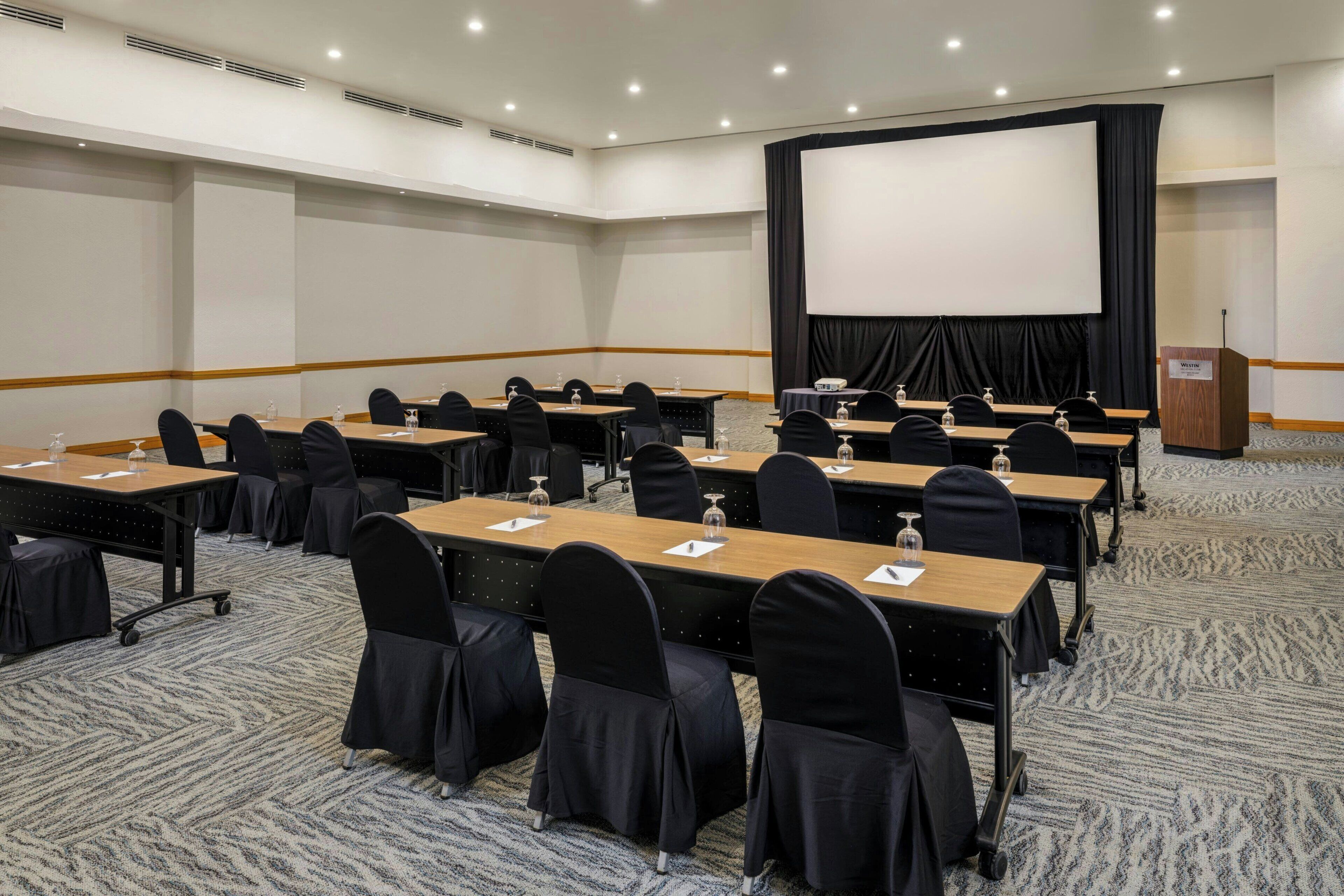 A conference room with multiple tables, chairs, a podium, and a large screen.