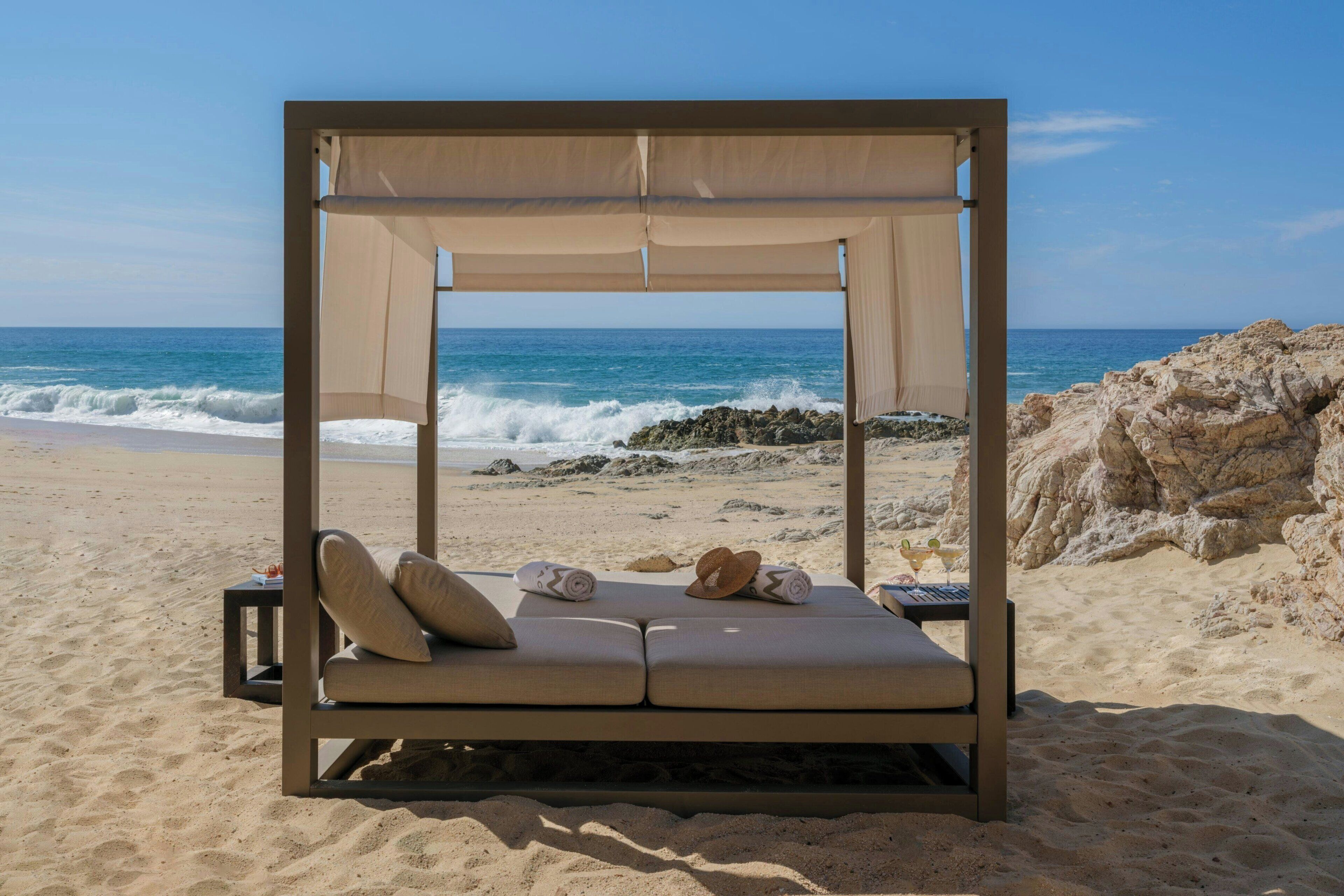 A beach cabana with a canopy, two cushioned benches, a hat, and a bottle of water.
