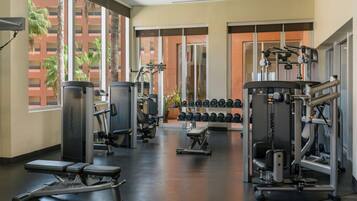 Ein Fitnessstudio mit verschiedenen Trainingsgeräten, darunter Kraftgeräte und Kurzhanteln, große Fenster und Blick auf Palmen.