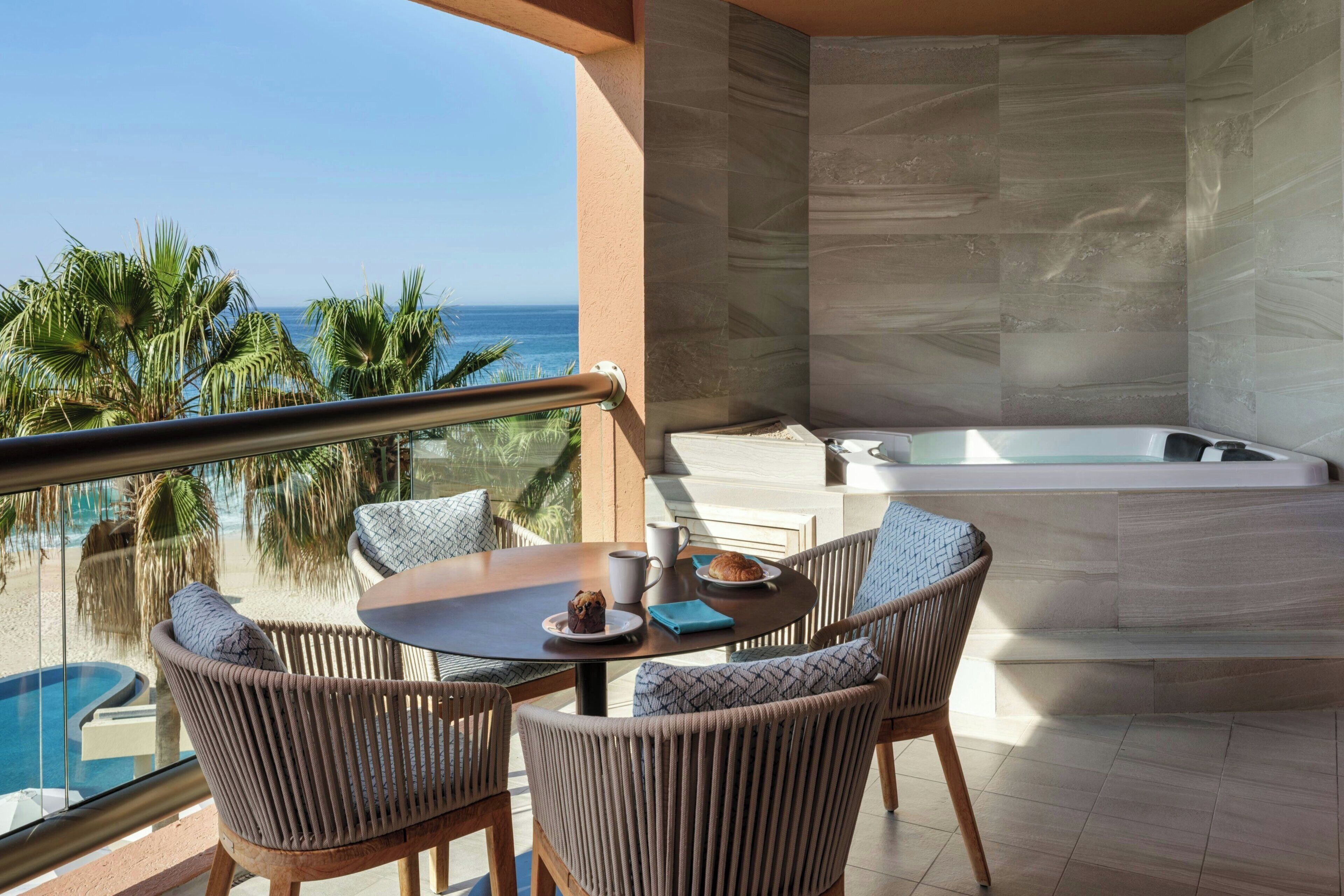 A balcony with a view of the ocean, a table with a muffin and a cup, and a jacuzzi.