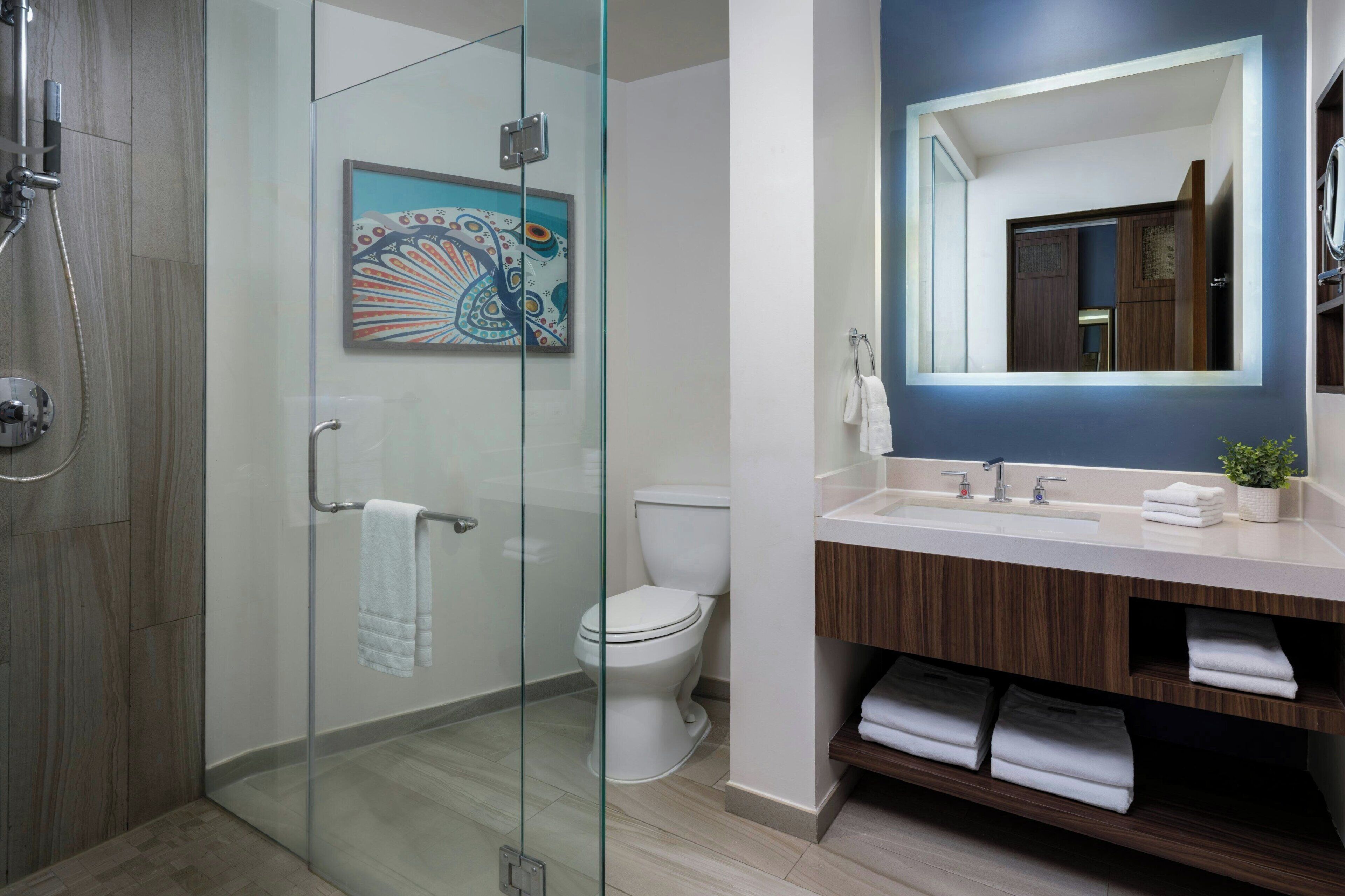 A modern bathroom with a glass shower enclosure, a white toilet, a wooden vanity with a mirror, and a colorful artwork on the wall.