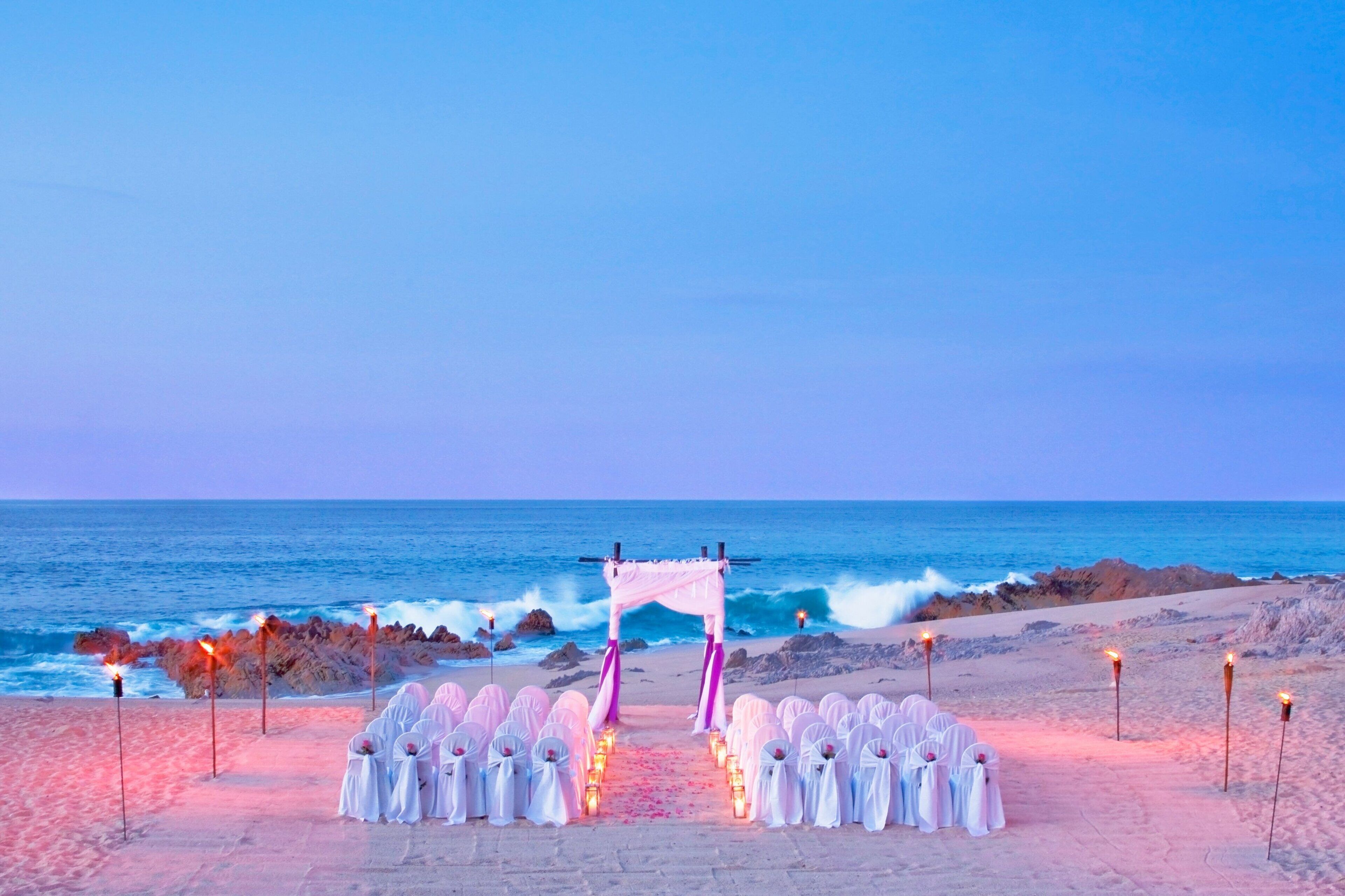 An outdoor wedding ceremony setup on a beach with a white archway, guests seated on chairs, and torches lining the aisle.