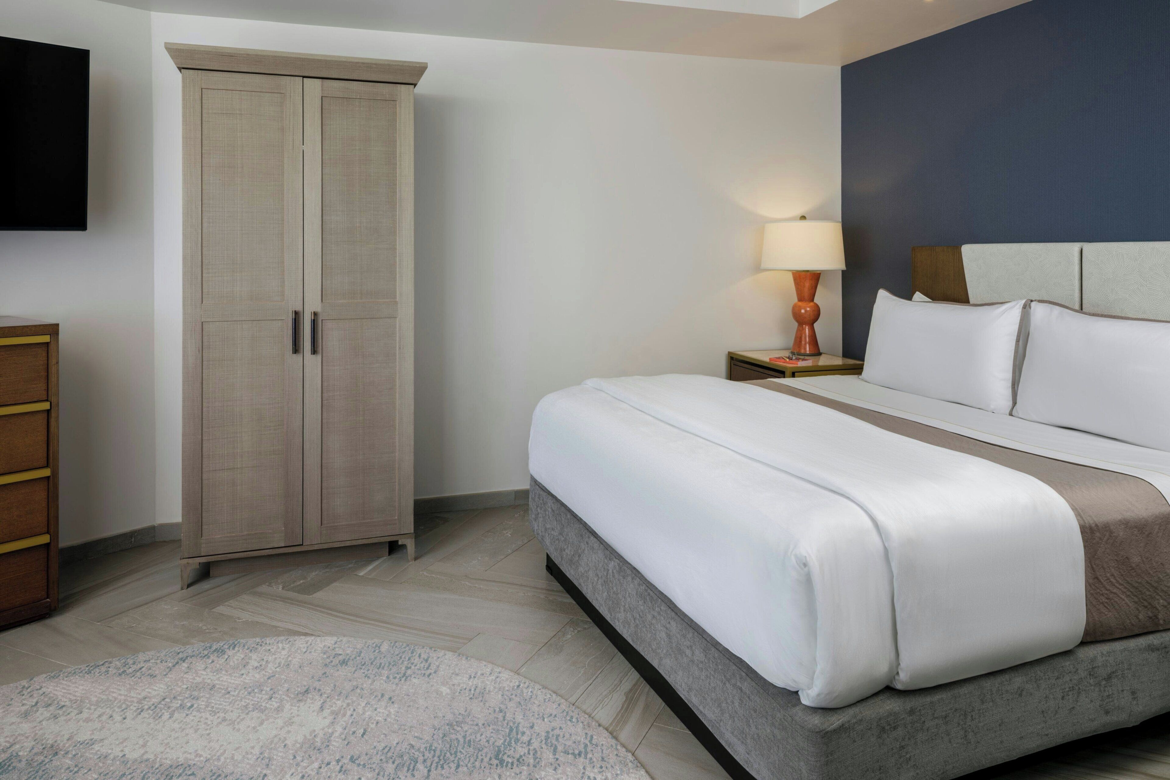A hotel room with a bed, a wardrobe, a nightstand with a lamp, and a television on the wall.