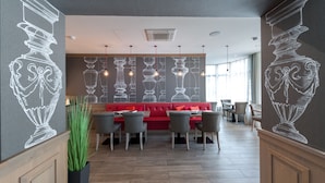 Un restaurante con una decoración moderna, que cuenta con un sofá rojo, sillas grises y una pared con ilustraciones de jarrones blancos.