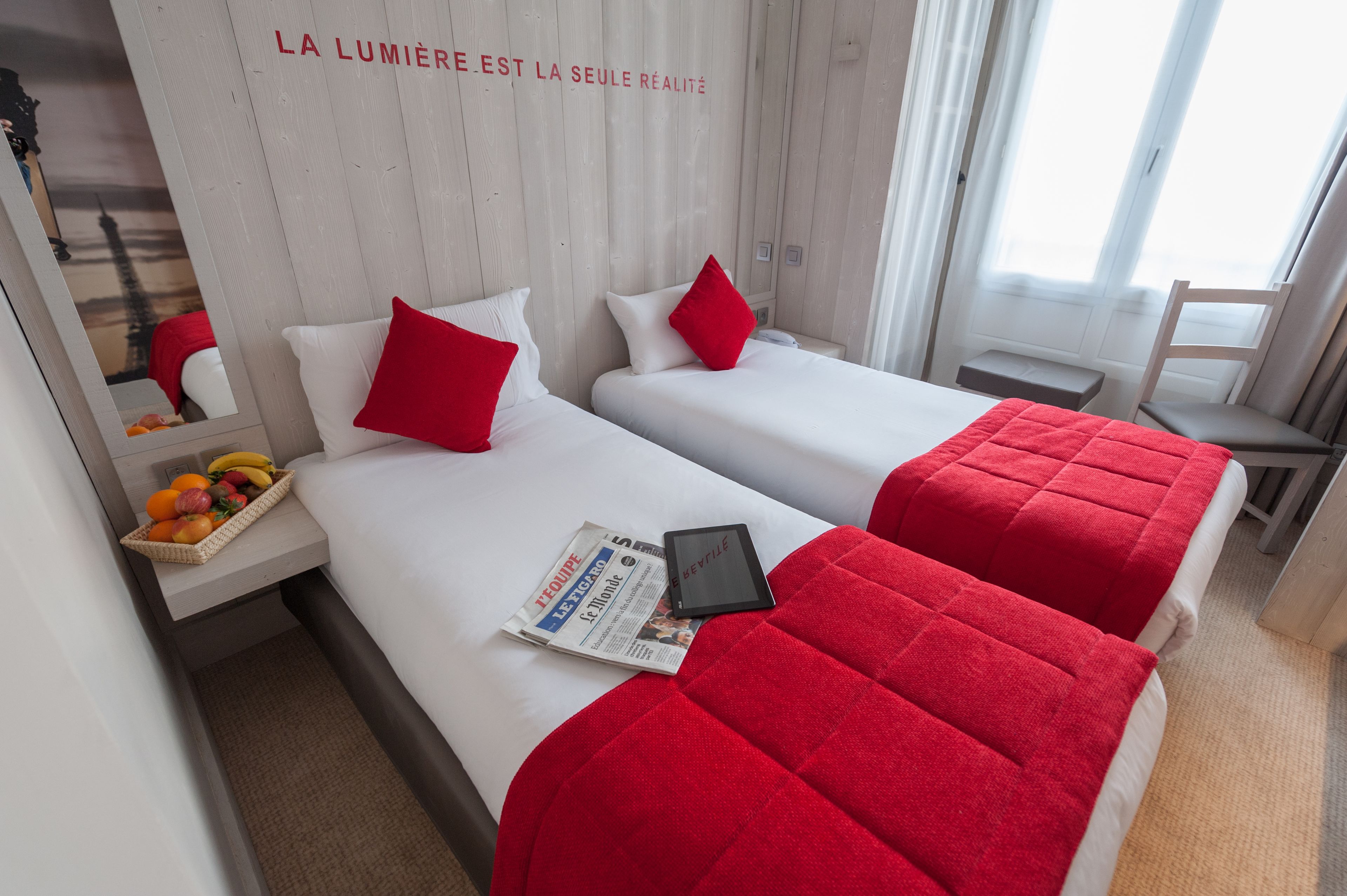 A hotel room with two beds, each with a red blanket and pillow, a small table with a fruit basket, and a newspaper.