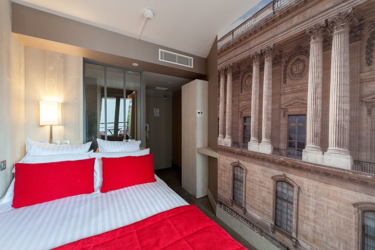 Une chambre d’hôtel avec un grand lit, une literie rouge et blanche, un miroir et une fresque représentant une architecture classique sur le mur.
