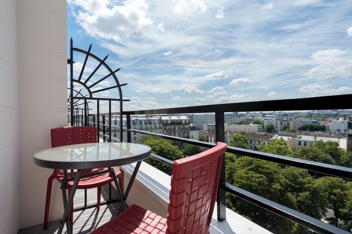Un balcon avec une table en verre et des chaises rouges, donnant sur un paysage urbain avec des immeubles et des arbres.