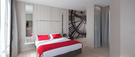 A modern hotel room with a large bed, a mirror, a clock mural, and a view of the city.