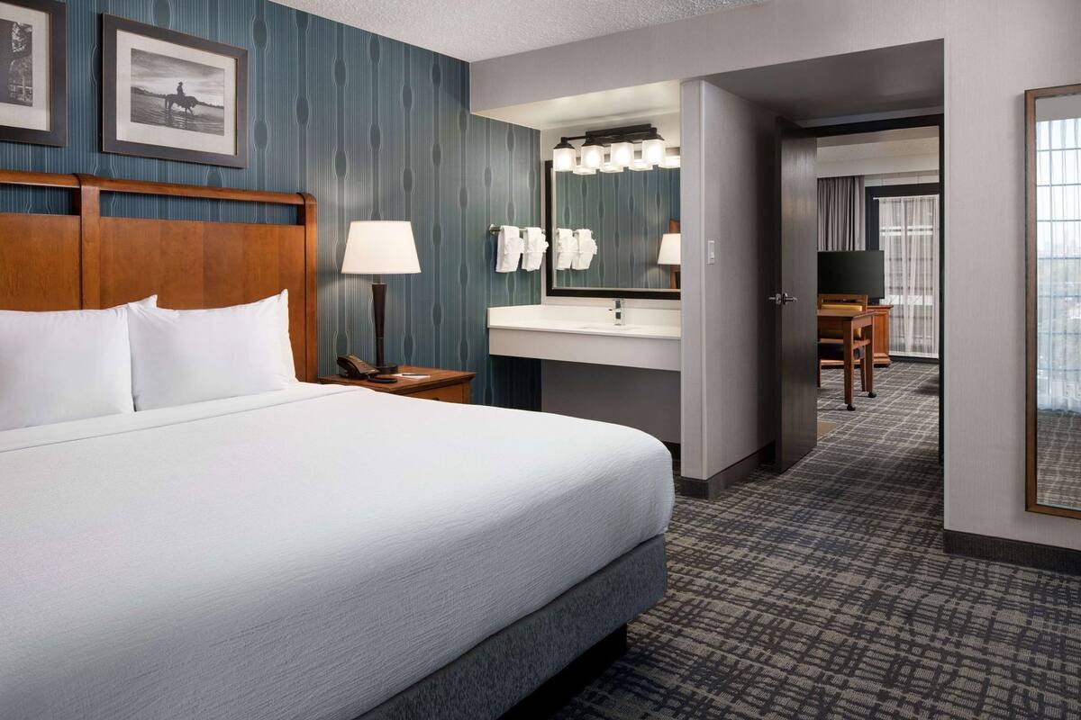 A hotel room with a bed, a bedside table with a lamp, a mirror, a bathroom with a sink and towel rack, and a view of the hallway.