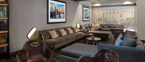 A modern hotel lobby with a sectional sofa, leather chairs, and a coffee table. There are framed pictures on the wall, a bookshelf, and a large window with blinds.