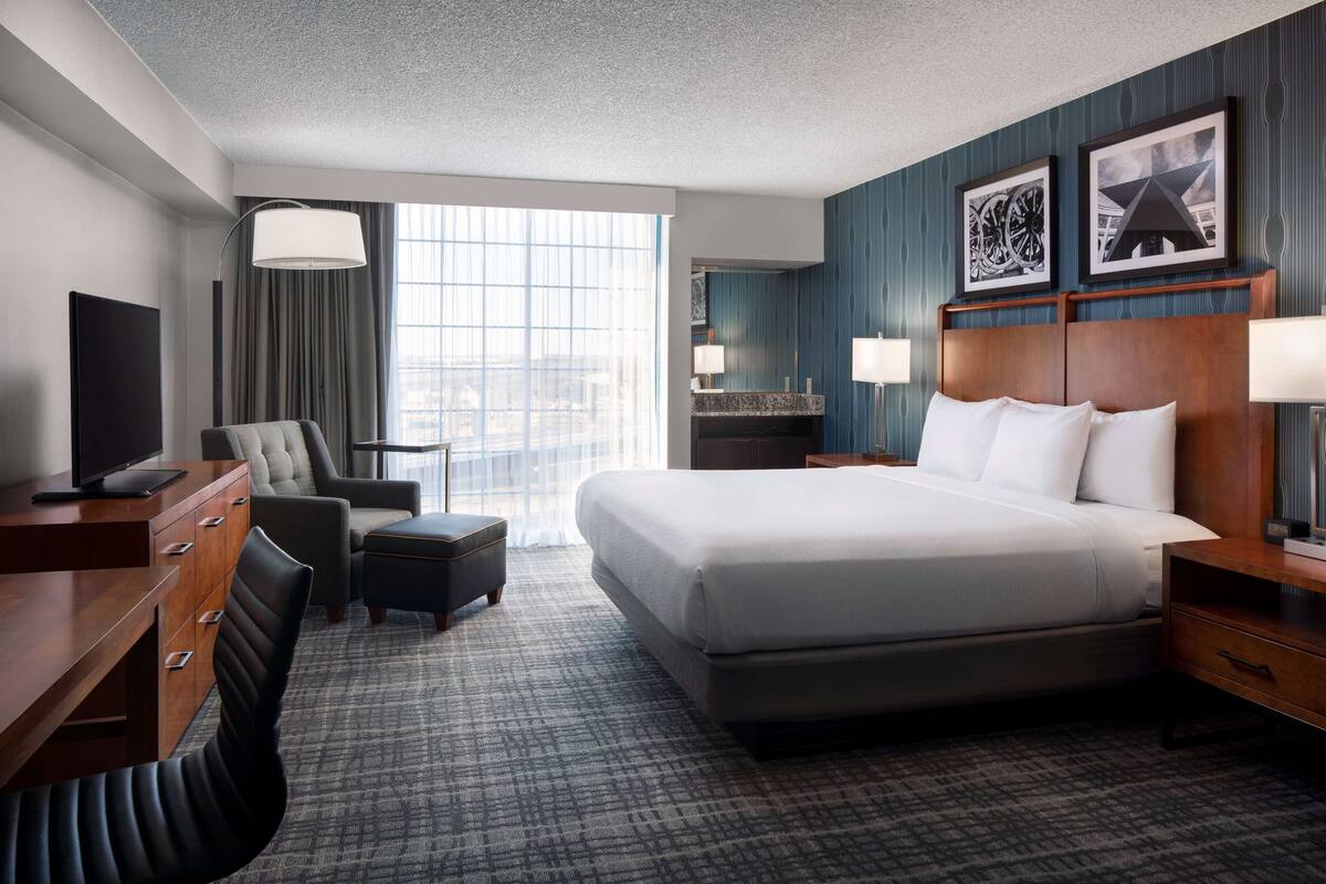 A hotel room with a large bed, a desk with a TV, a chair, a sofa, and a window with curtains.