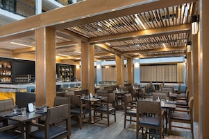 A modern restaurant with wooden beams, multiple dining areas, and a bar with a large screen.
