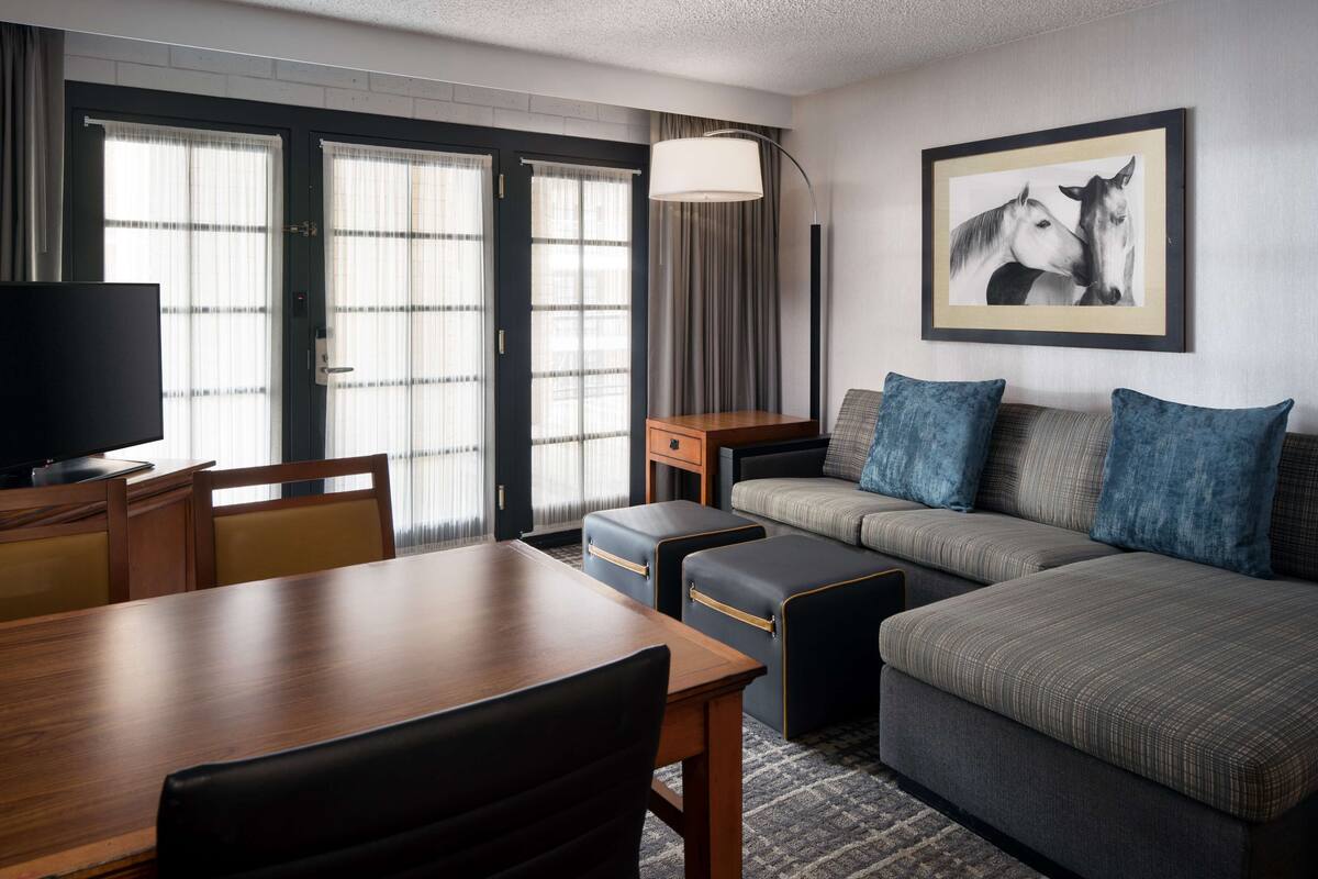 A hotel room with a wooden dining table, a sofa with blue cushions, a TV, and a framed picture of horses on the wall.