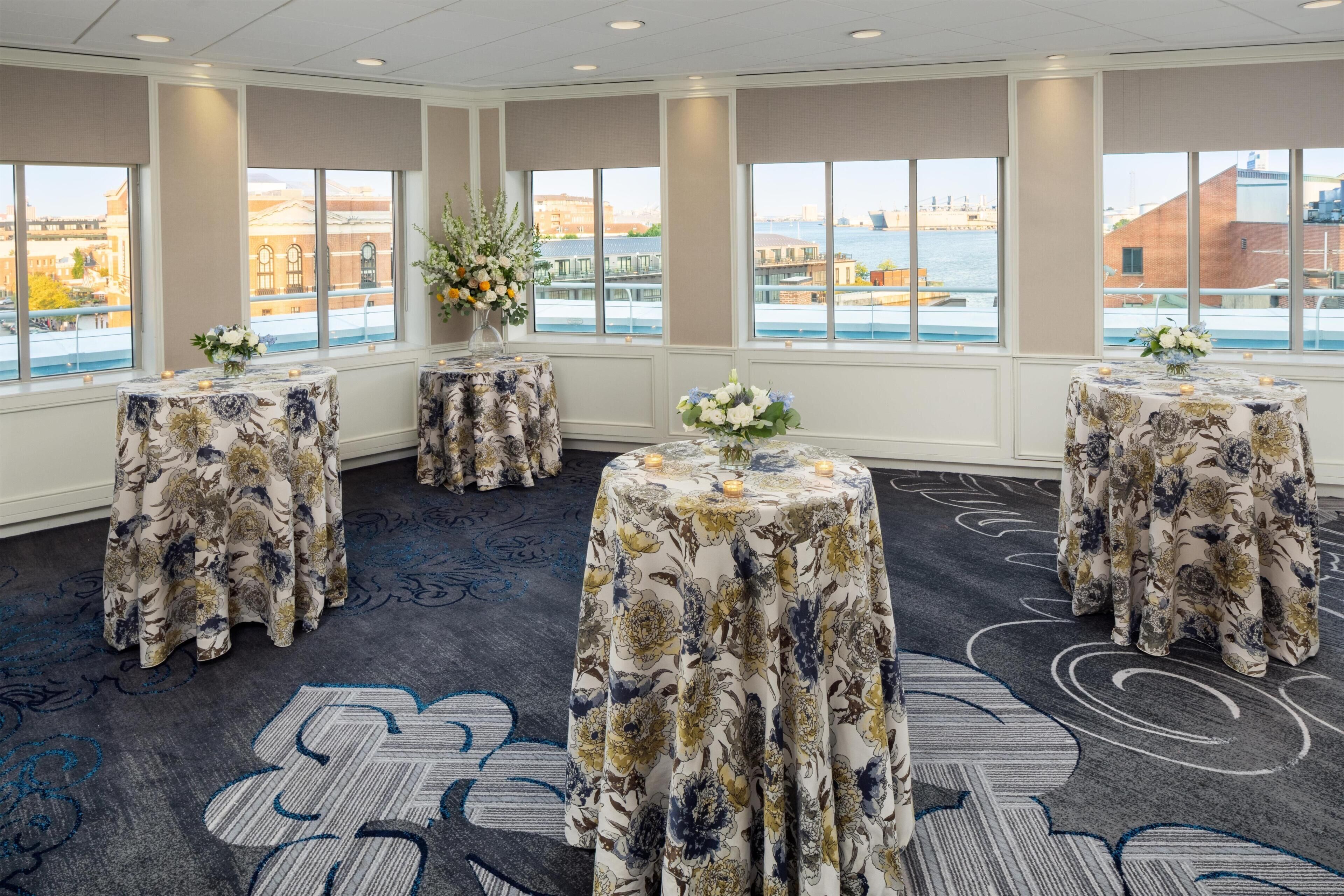 Ein Hotelzimmer mit großen Fenstern, Tischdecken mit Blumenmuster und einem Teppich mit geometrischem Design.