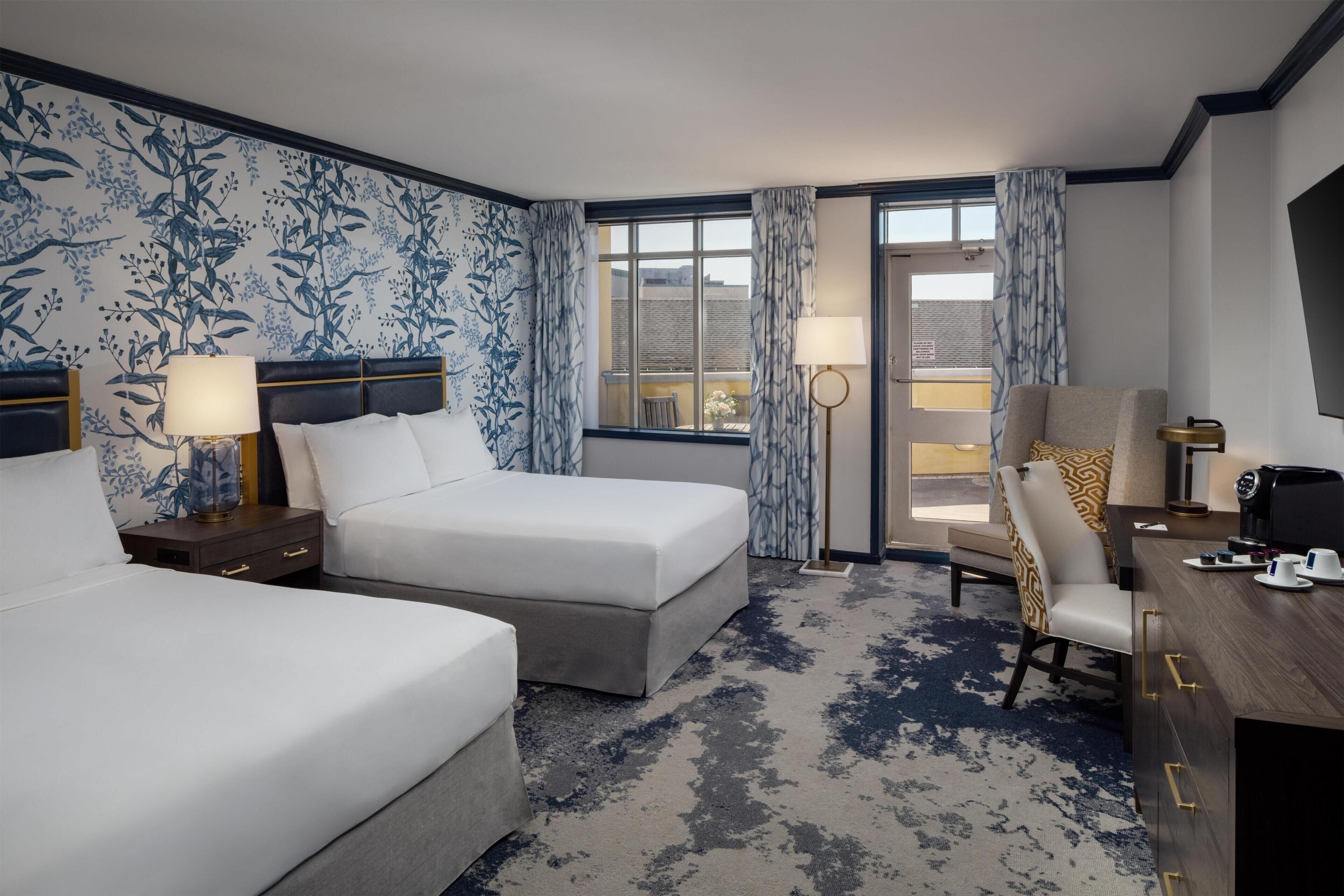 A hotel room with two beds, a desk, a chair, a coffee maker, and a view of the city.