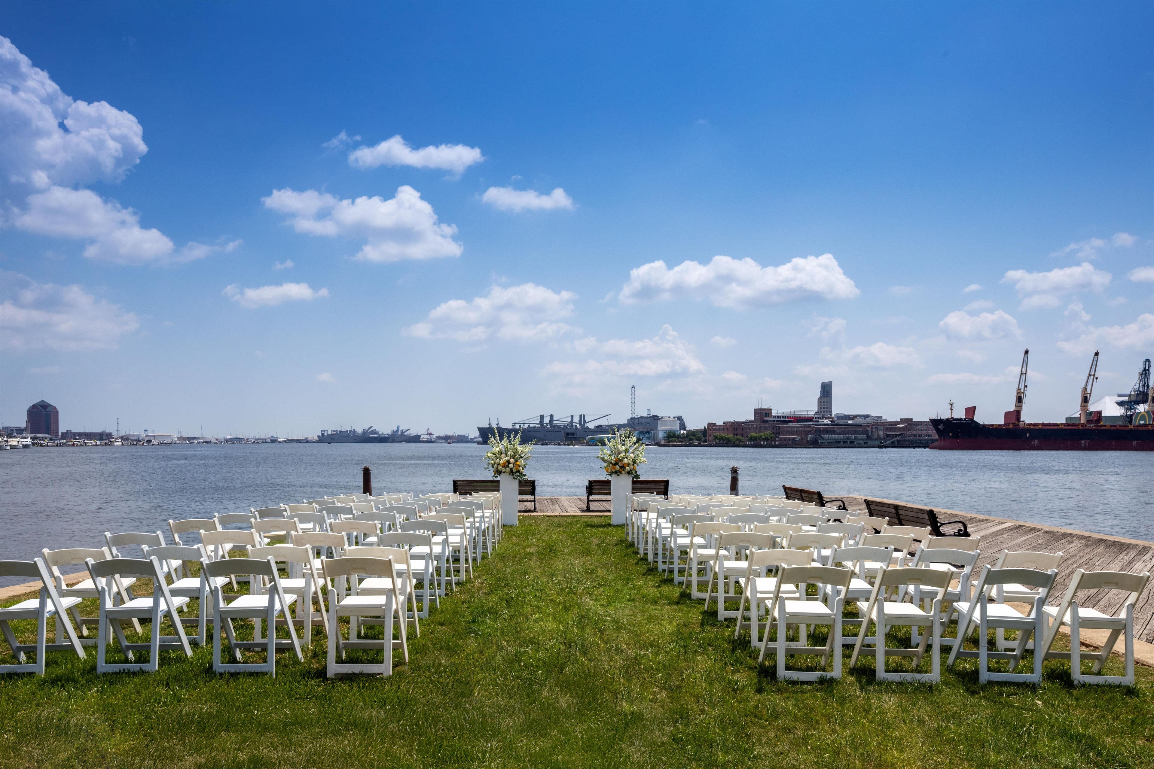 A waterfront venue with rows of white chairs arranged for an event, a grassy foreground, and a clear view of the water and distant buildings.