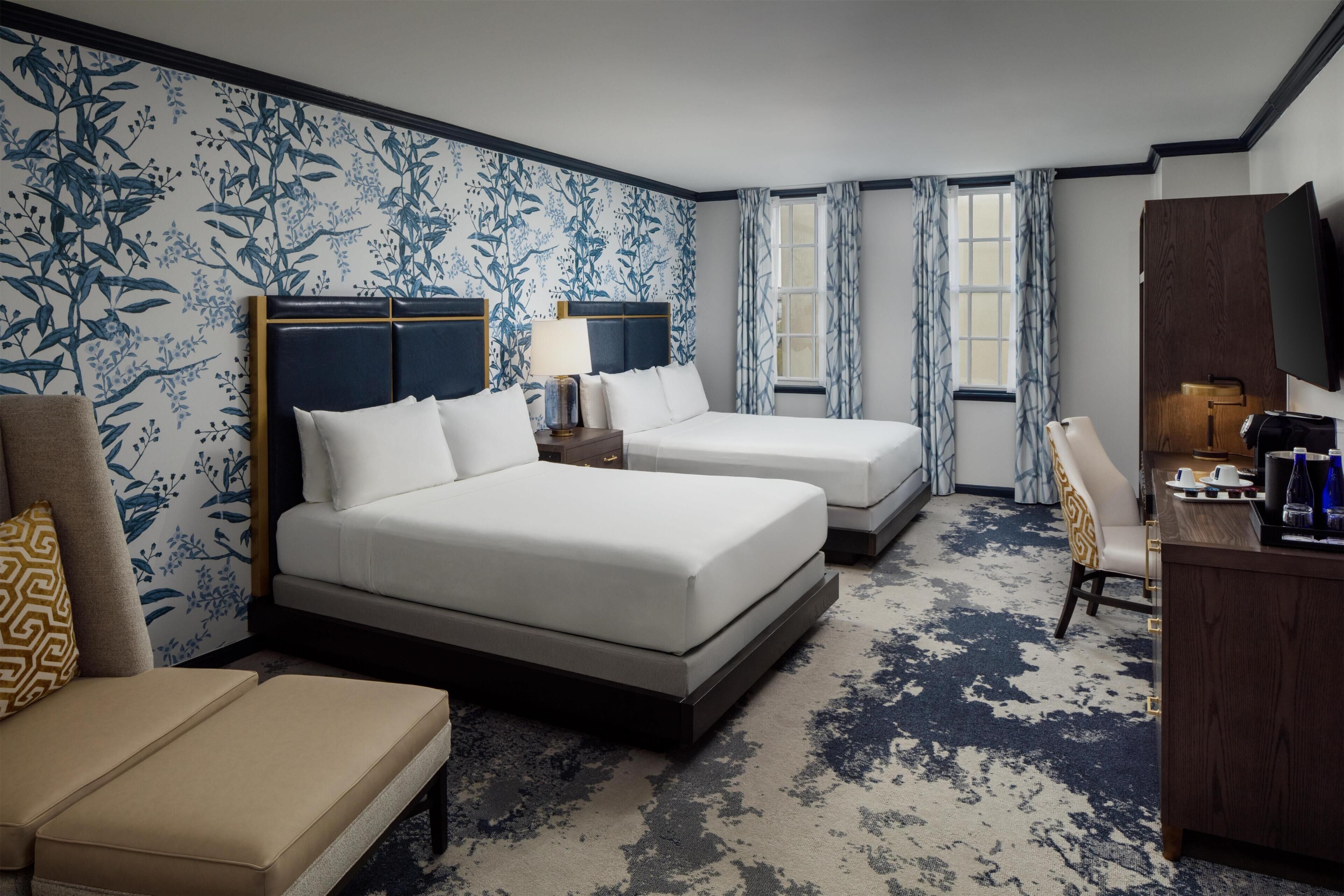 A hotel room with two beds, a desk, a chair, a TV, and a window with curtains.