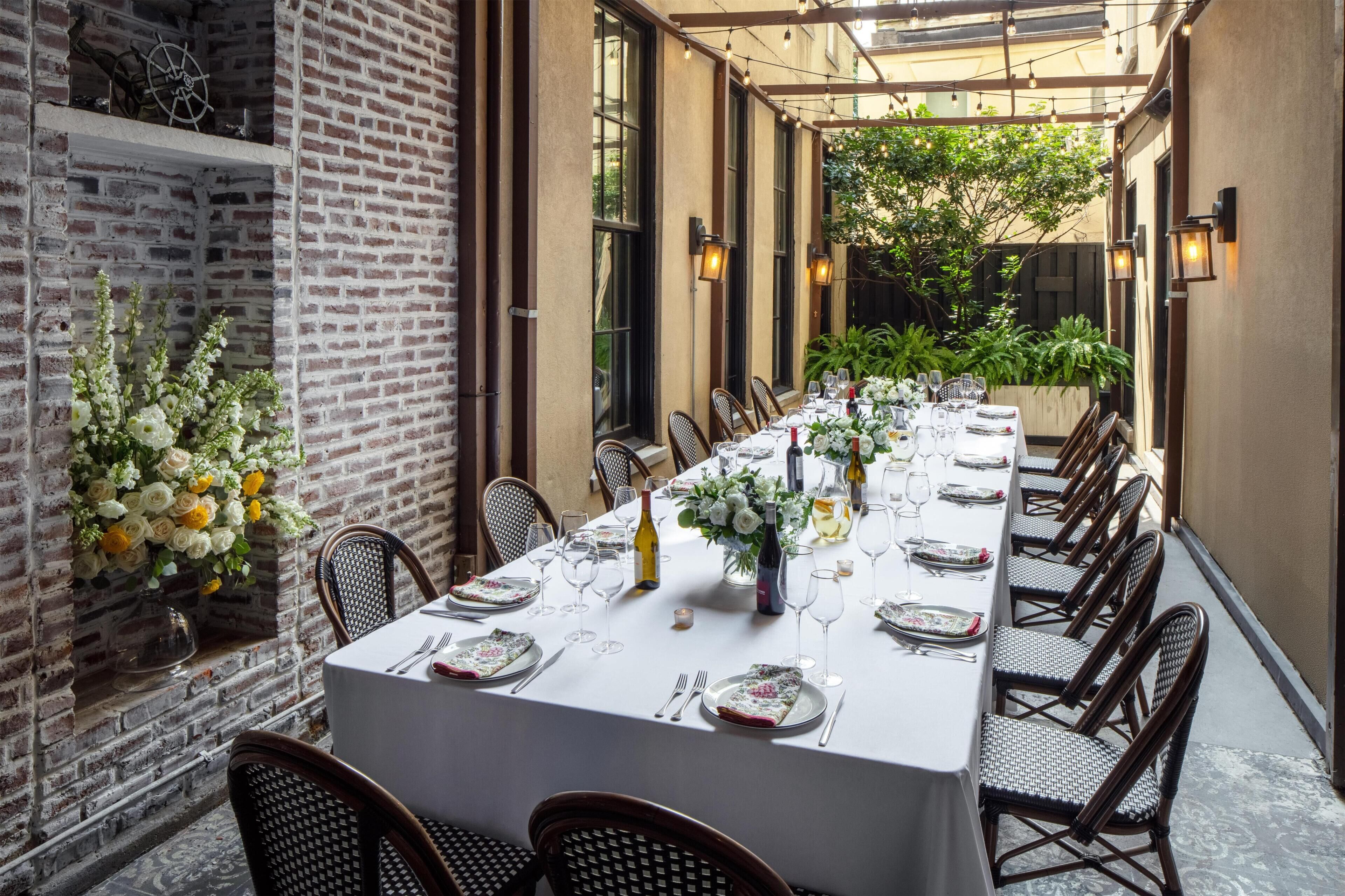 A long dining table set for a meal with chairs on both sides, in a covered outdoor area with brick walls and potted plants.