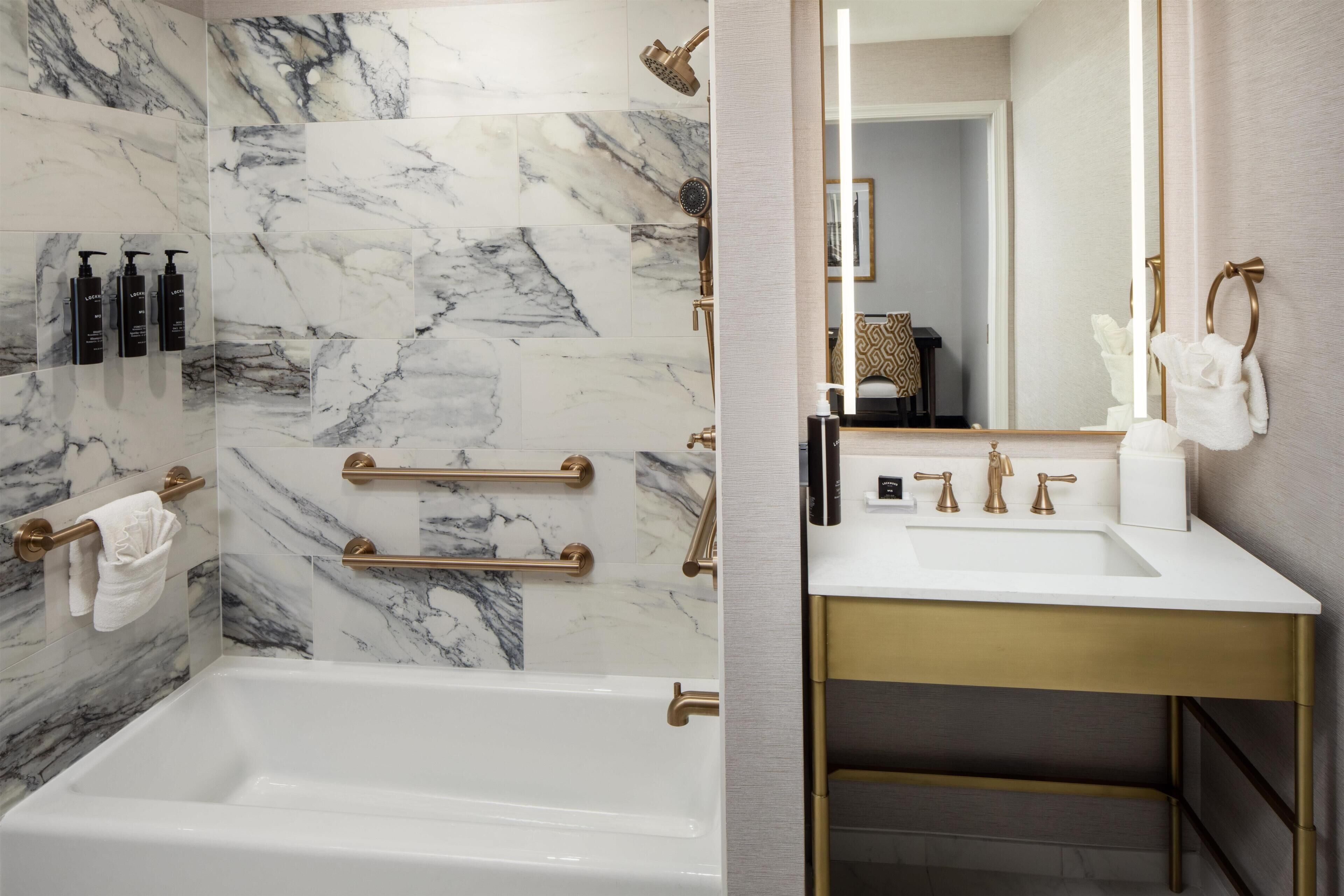A bathroom with a marble wall, a bathtub, a gold-colored towel bar, and a mirror with a towel hanging.