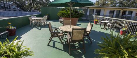 A rooftop patio with a round table and chairs, surrounded by potted plants and a green umbrella.